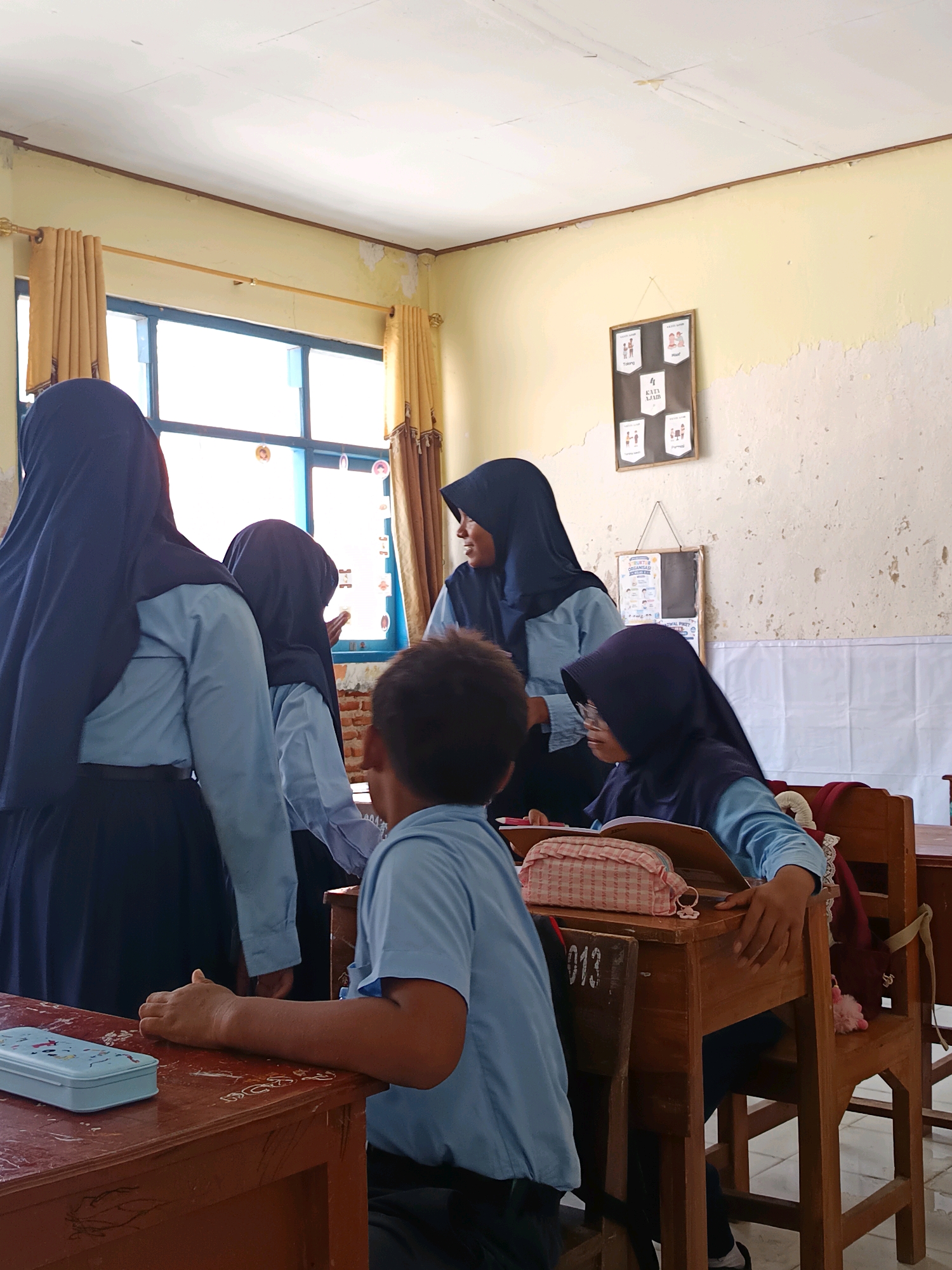 In a classroom in Labuhan, Indonesia, five students are visible. Four of the students are wearing light blue shirts and dark blue headscarves, suggesting they are female and dressed in school uniforms. One student is wearing a similar light blue shirt but does not have a headscarf. The students appear to be engaged in an activity at their desks, with two seated and three standing or leaning in. One student is looking at an open book, and another is gesturing towards it. The classroom environment is simple, with wooden desks and chairs, yellow-painted walls that show some signs of wear, and a window with light brown curtains. There are also some framed posters on the wall and a whiteboard behind the students. The overall impression is of a typical classroom setting in a school.