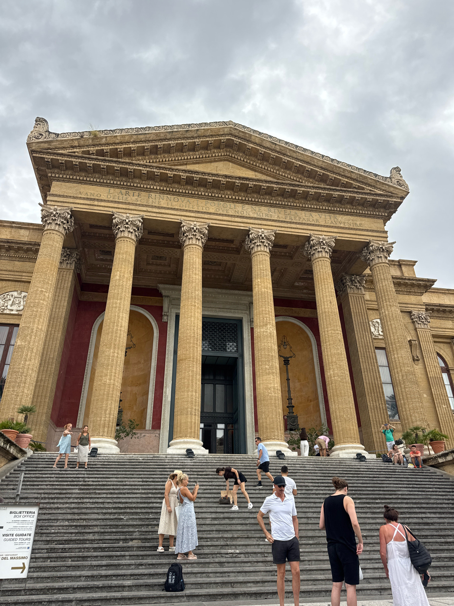 The image shows the exterior of the Teatro Massimo in Palermo, Italy, a grand opera house with a prominent Neoclassical facade. A wide, imposing staircase leads up to the entrance, which is flanked by massive Corinthian columns. The sky is overcast, casting a diffused light on the scene. Several people are scattered across the steps, some posing for photos, others conversing, and a few are looking at the architecture. One person is taking a photo of another woman. A signpost on the left side of the stairs indicates "BIGLIETTERIA BOX OFFICE" and "VISITE GUIDATE GUIDED TOURS." The overall atmosphere is one of a popular tourist destination on a cloudy day.