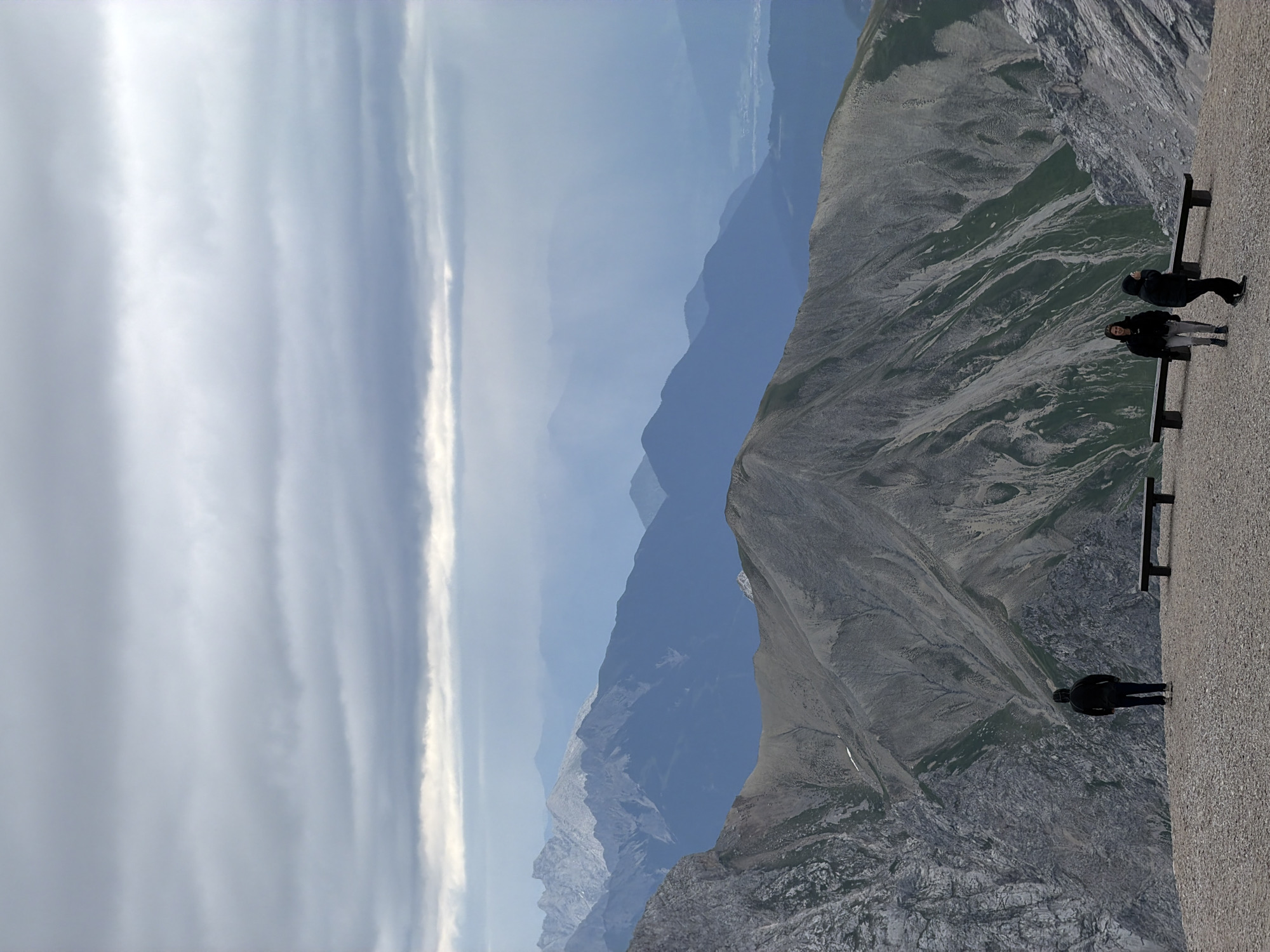 The image depicts a panoramic view of a mountainous landscape, likely from a high vantage point, potentially the Zugspitze, Germany's highest peak, near Garmisch-Partenkirchen. In the foreground, a gravelly, rocky terrain is visible, with three individuals present. One person stands alone facing away from the camera, towards the vast mountain range. To the right, two other individuals, dressed in dark clothing, are walking. Two dark wooden benches are positioned on the gravel area. The middle ground is dominated by rugged, grey mountains with visible striations and patches of green vegetation. The background is a series of layered mountain ranges fading into a hazy blue, with a dramatic, cloudy sky overhead. The clouds are dense and layered, creating a somber and atmospheric effect. The overall scene conveys a sense of natural grandeur and isolation.