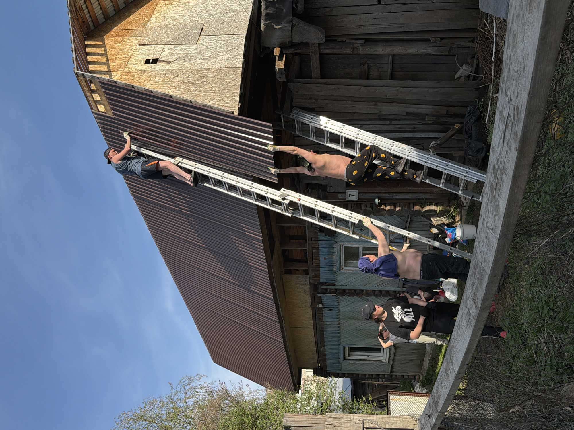 A group of individuals is engaged in construction work on a building with metal roofing and wooden siding, potentially located in Perm, Russia. One man is on a ladder, actively working on the roofing. Another man is lying across two ladders, seemingly as part of a support or holding action. Below, other men are standing on the ground, possibly assisting or observing the process. The overall setting appears to be a rural or suburban area. The sky is blue with a few clouds. The building is old, made out of wood.