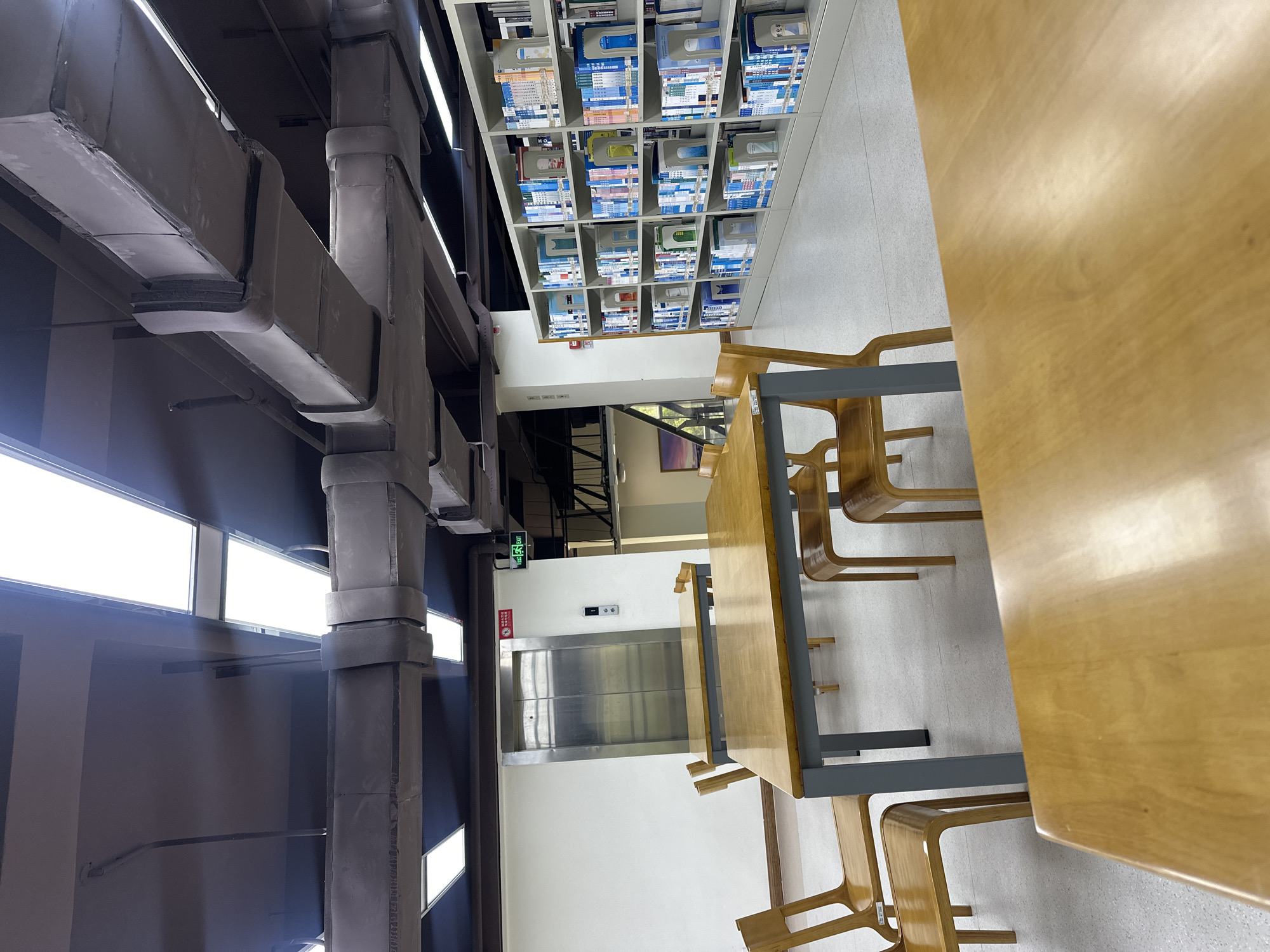 The image depicts the interior of a building, possibly a library or study area in the Chongqing High-tech Zone, China. There are tables and chairs arranged, suggesting a space for reading or working. Shelves filled with files or documents line one wall. Visible overhead are industrial-style air ducts. An elevator is situated in the center, adjacent to the tables and chairs. Windows are present on one side, providing natural light.