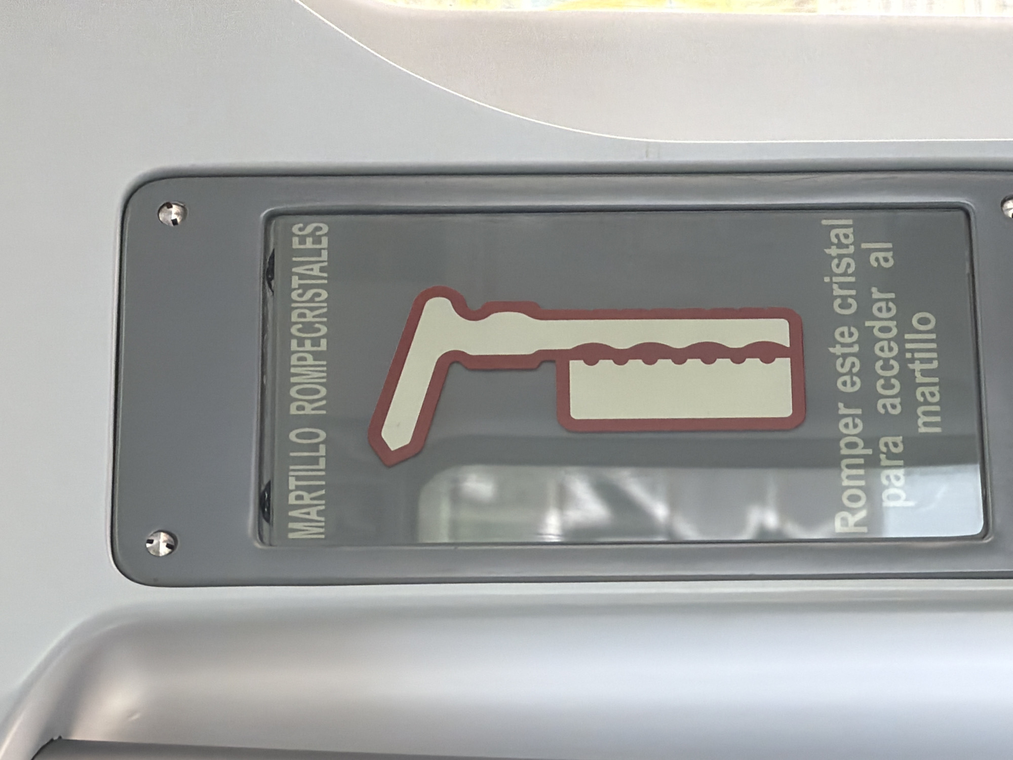 The image shows a sign in Spanish that reads "MARTILLO ROMPECRISTALES" and "Romper este cristal para acceder al martillo" with an image of a hammer. The sign is likely located in a vehicle, possibly a bus or train. There is no verifiable evidence in the image to suggest Madrid, Spain.