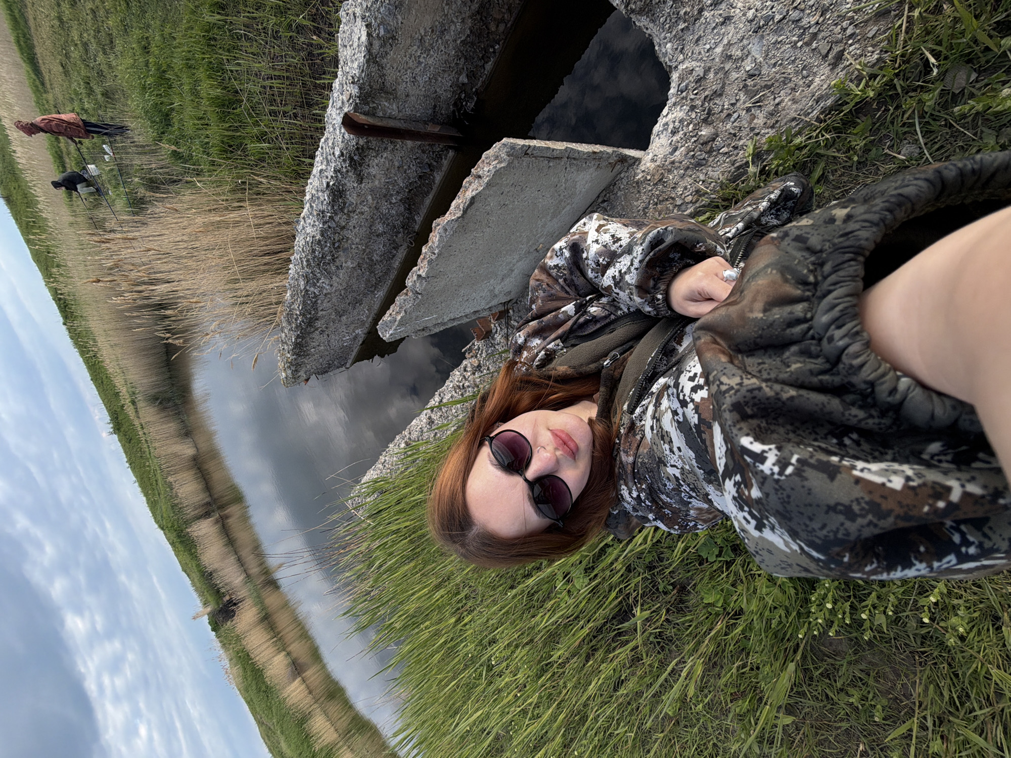 A woman is lying on the grass near a concrete structure, likely part of a waterway or canal. She is wearing a camouflage jacket and sunglasses. The location appears to be in a rural area with tall grass and water. Two people are fishing in the distance. The sky is cloudy. The location appears to be Novomargaritovo, Russia.