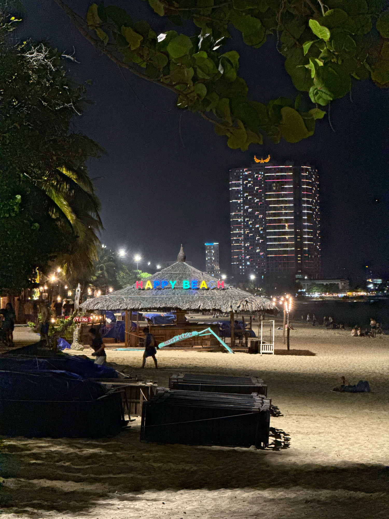 The image depicts a nighttime scene on a beach in Nha Trang, Vietnam. In the foreground, folded black tarps or covers are stacked on the sand. Further back, a thatched-roof structure, possibly a beach bar or cafe, is illuminated by colorful neon lights that spell out "HAPPY BEACH." A few people are visible near this structure, some walking and others standing. To the left, palm trees are present, with string lights illuminating the area. In the background, a tall, modern building is brightly lit with horizontal lines of colored lights. Other smaller buildings and lights indicating a city skyline are visible in the distance. The beach itself is sandy, with some scattered beachgoers in the mid-ground and background. The overall atmosphere suggests a lively evening at a coastal location.