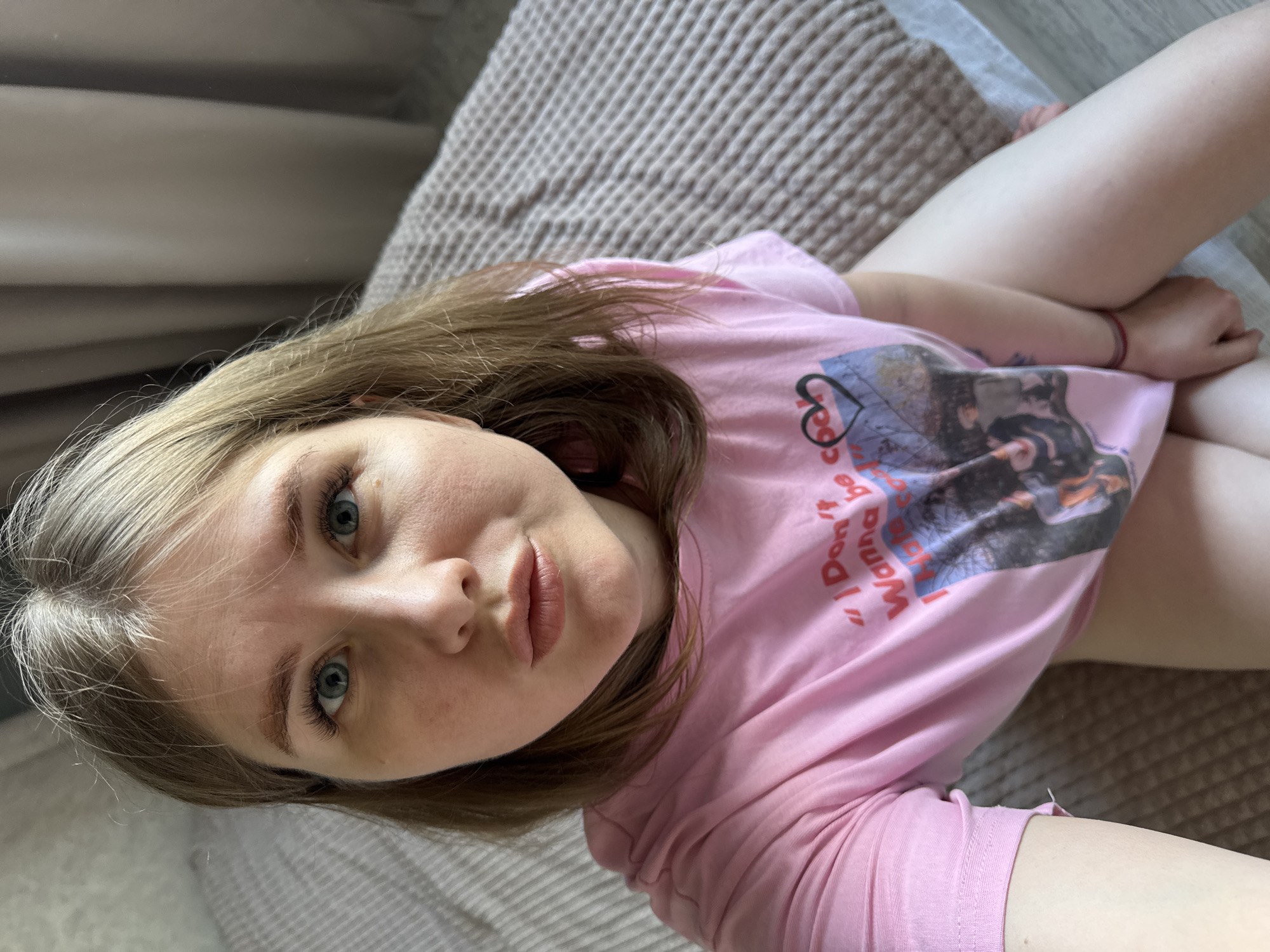 This image depicts a fair-skinned adult female, located indoors in Gornyi (Dvurechenskoe S/P), Russia. She possesses light brown/blonde hair and blue eyes with enhanced eyelashes, gazing directly at the camera with slightly pursed lips. She is wearing a short-sleeved pink t-shirt featuring a graphic print with the visible text "I Don't Wanna be cooL" and a partial image below it, including a black heart outline. A thin red bracelet is present on her right wrist. Her bare legs are visible below the shirt, with her hands resting on her thighs. She is seated or reclining on a bed or sofa covered with a textured, light beige blanket. The background on the left consists of a plain light-colored wall, while the right side features beige curtains. A light wood-patterned floor is visible in the lower right.