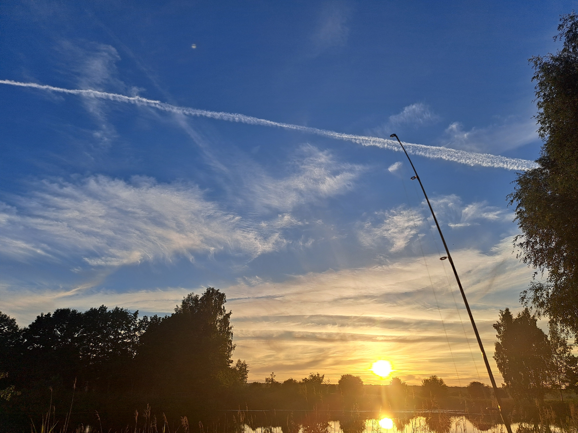 The image captures a serene outdoor scene at dawn or dusk, likely near Riga, Latvia. The sun, a bright orb, is low on the horizon, casting a warm golden glow across the sky and reflecting on the calm surface of a body of water. Wispy clouds stretch across the deep blue sky, with a prominent contrail from an aircraft streaking horizontally.
In the foreground, the silhouette of a fishing rod stands upright, angled towards the sky, suggesting the activity of fishing. Reeds and tall grasses line the bank of the water, adding to the natural setting. The opposite bank is lined with the dark silhouettes of trees, creating a natural border to the landscape. The overall mood is peaceful and tranquil, evoking a sense of calm and connection with nature. The time of day is clearly indicated by the low sun and the soft lighting. There is no visible text in the image.
