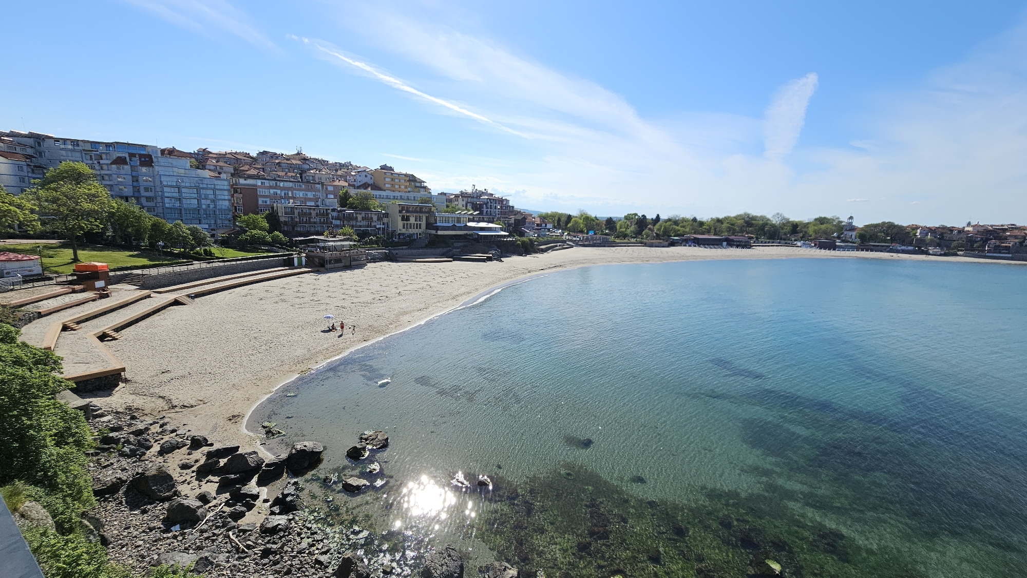 The image shows a beach in Sozopol, Bulgaria. The beach has light beige sand with a few people and a sun umbrella visible near the waterline. The water is clear and turquoise, revealing rocks and seaweed on the seabed. The foreground shows dark rocks covered in what appears to be vegetation.
In the background, there is a line of buildings and trees along the coast. The buildings vary in height and architectural style. To the left of the beach, there's a series of stepped platforms leading down to the sand, with a small red-roofed structure. The sky is clear blue with some wispy clouds. The picture is taken from an elevated position, providing a wide view of the beach and the surrounding landscape.