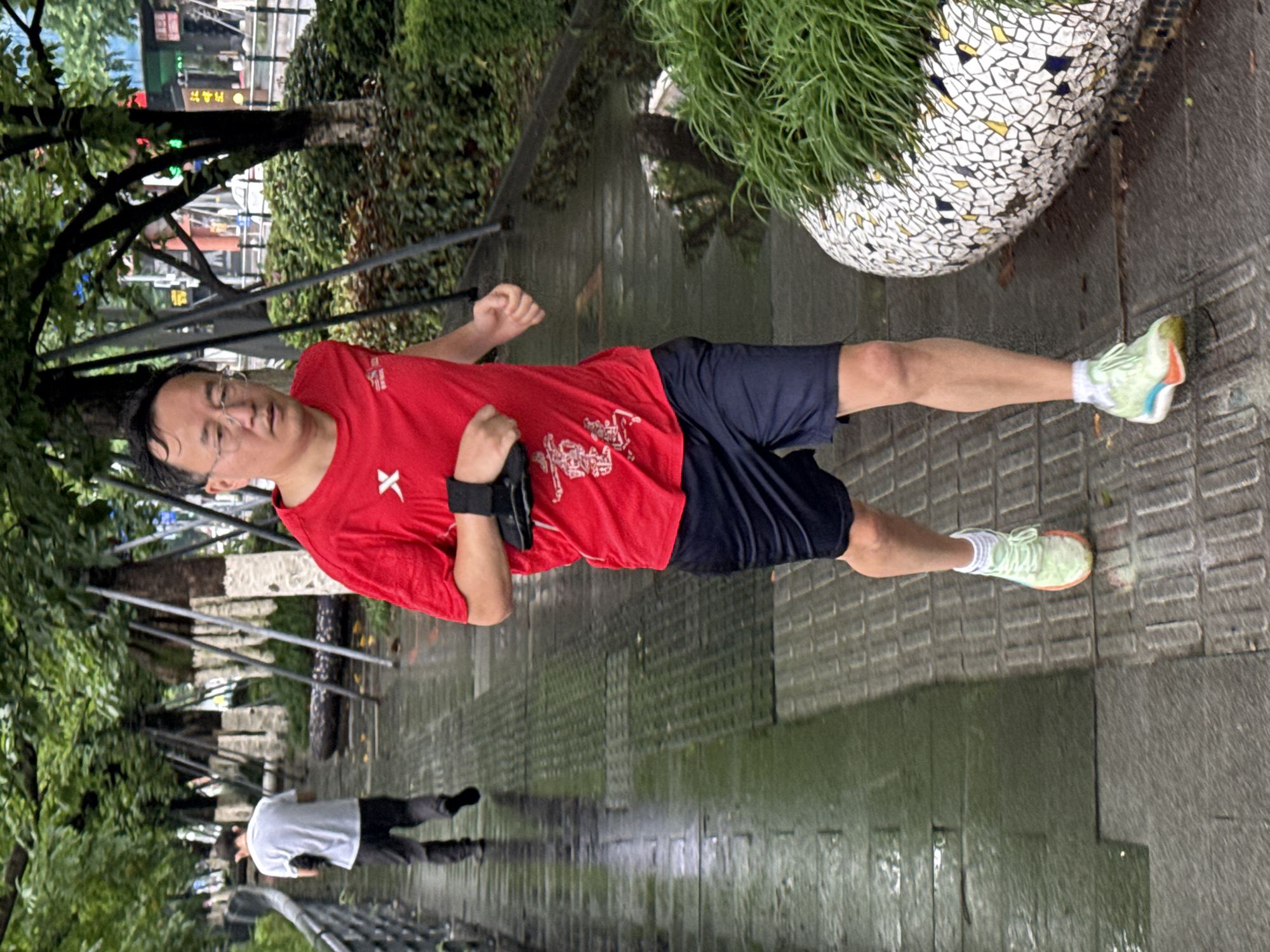 A man is jogging along a wet, tree-lined walkway in Nan'an District, China. He wears a red t-shirt, dark shorts, and athletic shoes. He has a black band around his upper arm. Another person is walking in the distance behind him. The area is likely a park or garden, with a tiled planter filled with foliage visible to the jogger's right.