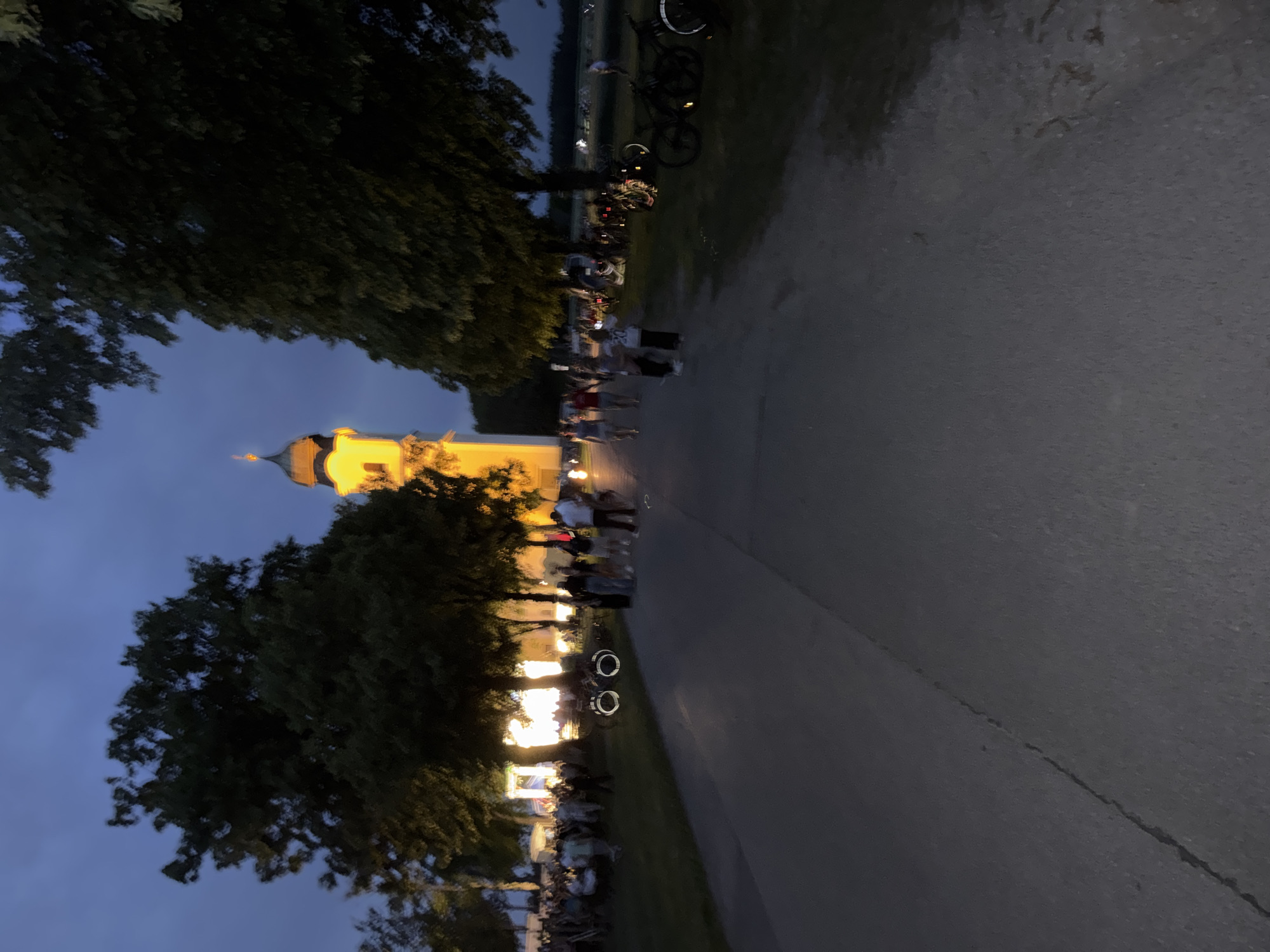 The image shows a twilight scene in Höhenkirchen-Siegertsbrunn, Germany, with a prominent illuminated church tower in the background. A pathway or road leads towards the tower, with trees lining the right side. Several people are walking on the pathway and gathered on the grass to the left, near illuminated tents or structures. Bicycles are parked on the grass to the right, near the trees. The sky is a deep blue, indicating dusk.