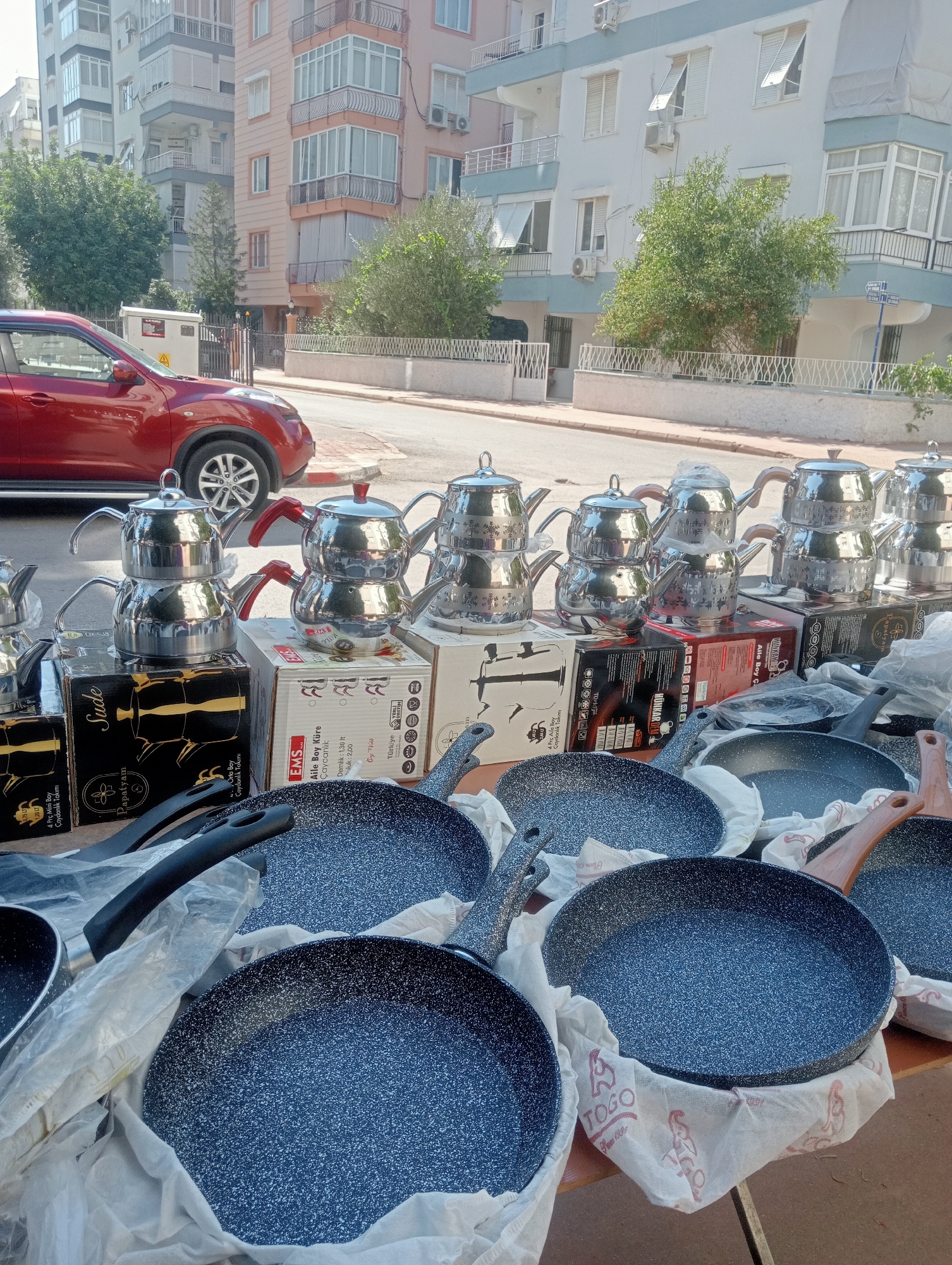 A street-level view on a sunny day in Antalya, Turkey, captures an outdoor stall displaying a variety of kitchenware. In the foreground, several new, dark-speckled frying pans are arranged on a table, some wrapped in white fabric. Behind the pans, stacked boxes contain tea sets. The tea sets are made of shiny metal, with traditional stacked designs, featuring spouts and handles in red or wood.
Further back, a red car is parked at the curb. The background is dominated by multi-story apartment buildings with balconies and windows. Green trees and bushes are visible alongside the buildings and on the street. A street sign is partially visible on the right side of the image. The overall scene suggests a commercial street or market area within the city.