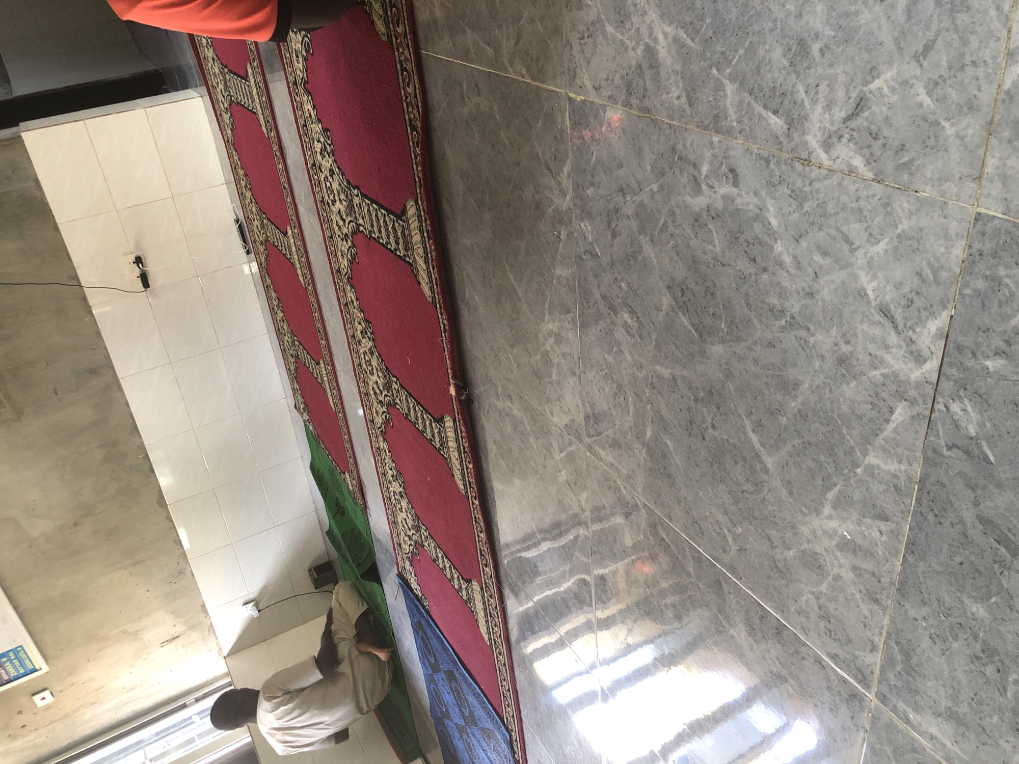 In Ado, Nigeria, a man wearing a light-colored robe is seated on a green mat, facing away from the camera. He is positioned on a prayer mat with a red and gold pattern, laid out on a gray tiled floor. Multiple similar prayer mats are visible in rows. To the left, a blue mat with a checkered pattern is also on the floor. The walls are tiled in white and grey. Electrical outlets are mounted on the wall, with one visibly connected to a charging cable. A sign with writing is affixed to the wall in the upper left corner. The overall scene depicts a prayer area, possibly within a mosque or a similar facility.