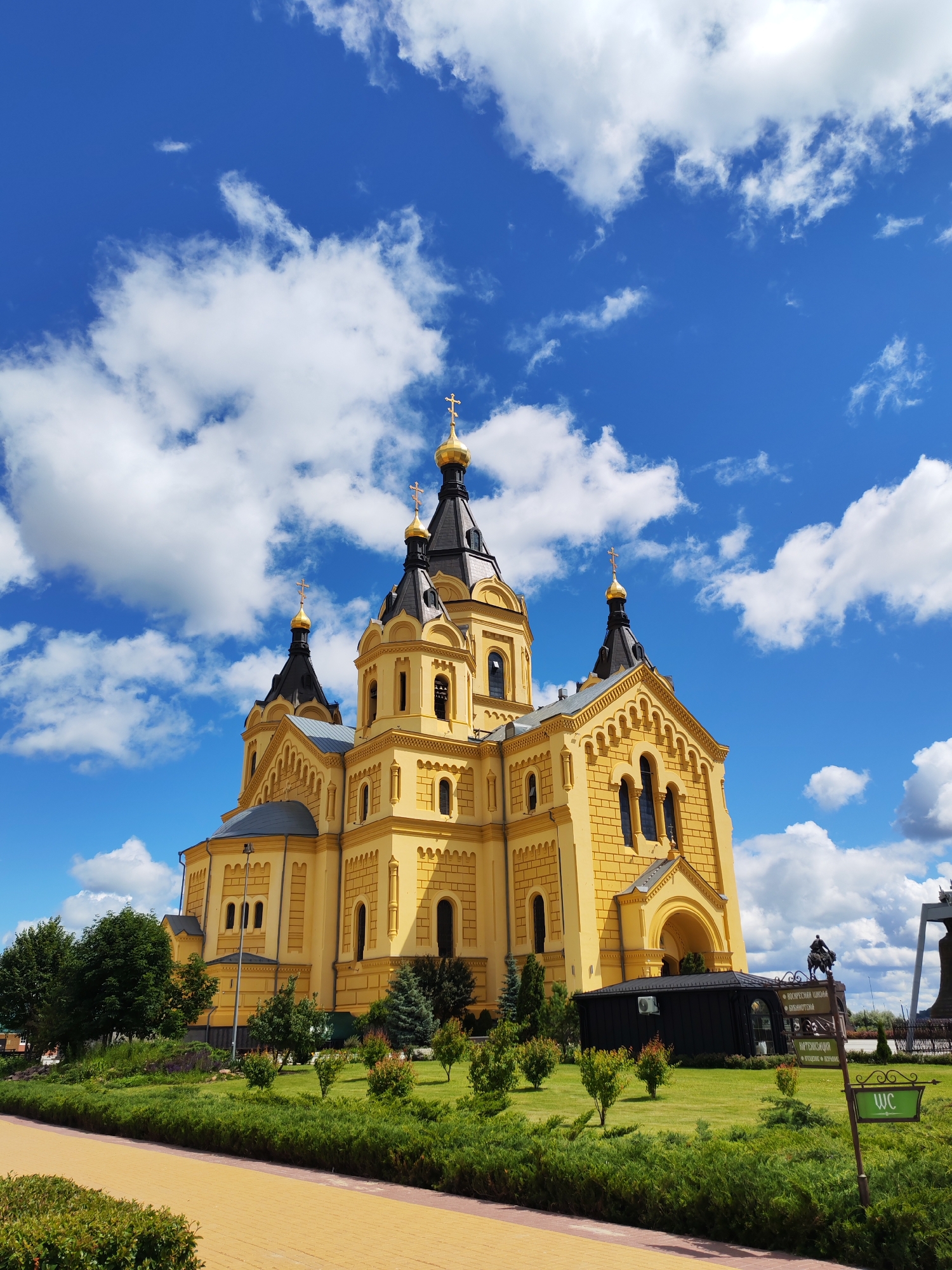 The image shows a bright yellow Orthodox church with multiple domes topped with gold crosses against a vibrant blue sky dotted with fluffy white clouds. The church features intricate architectural details, including arched windows and decorative friezes. In the foreground, a well-maintained lawn is bordered by neatly trimmed bushes. To the right of the church, there is a small black building, a signpost with text in Russian, a statue of a person on horseback, and a green sign indicating "WC". A yellow paved walkway is visible in the bottom left of the frame. The scene is bathed in daylight, suggesting it is daytime, and the clear sky with scattered clouds indicates fair weather. The overall impression is one of grandeur and peacefulness, likely in a public or historical site given the signage. The location is identified as Nizhny Novgorod, Russia.