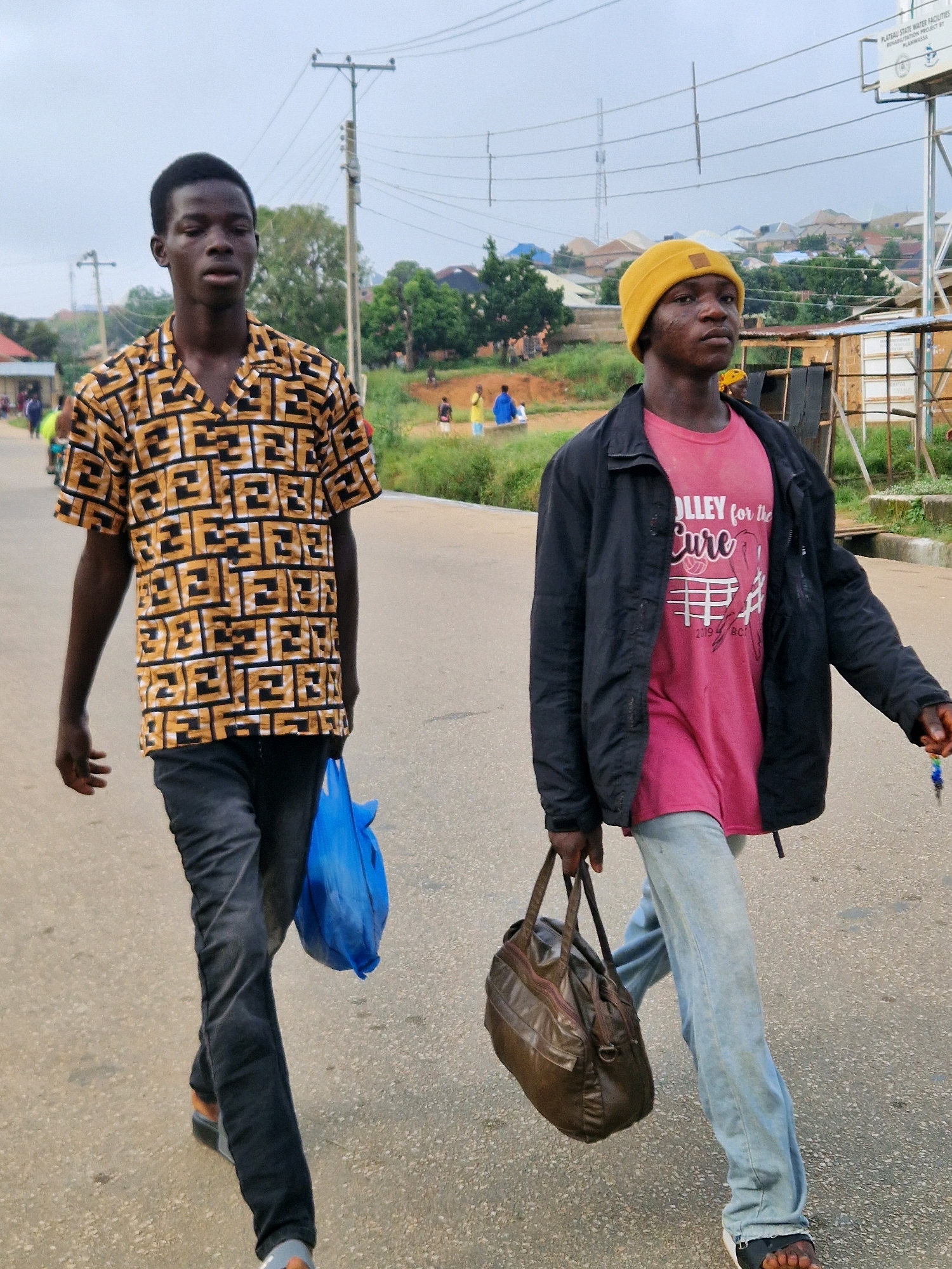 In Kabong, Nigeria, two young Black males walk along a paved road. The male on the left wears a short-sleeved shirt with a black and gold geometric pattern, dark pants, and dark sandals. He carries a bright blue plastic bag in his right hand. The male on the right wears a yellow beanie, a dark jacket over a pink t-shirt with graphic print, light blue jeans, and black and white sandals. He carries a brown leather satchel in his left hand. In the background, there are residential buildings, trees, utility poles, and a billboard. The sky is overcast.