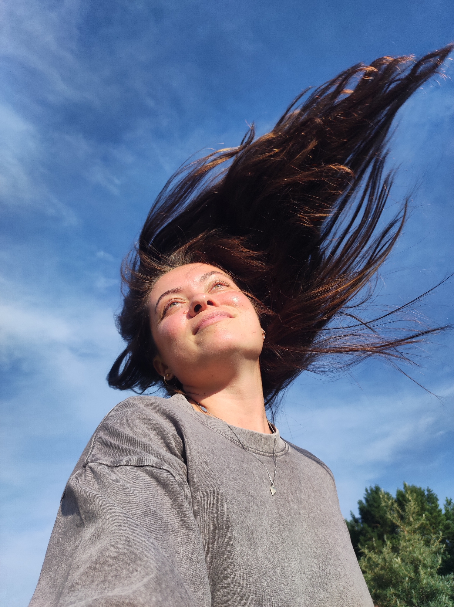 A low-angle, medium shot captures a young woman with long, dark hair being blown upwards by the wind against a bright blue sky with wispy white clouds. She is wearing a gray, long-sleeved t-shirt and a silver pendant necklace. Her head is tilted back, and she is looking up towards the sky with a serene expression, her face illuminated by sunlight. The background shows a blurred green tree line. This scene is not identifiable as being in the city of Фоки, Russia.