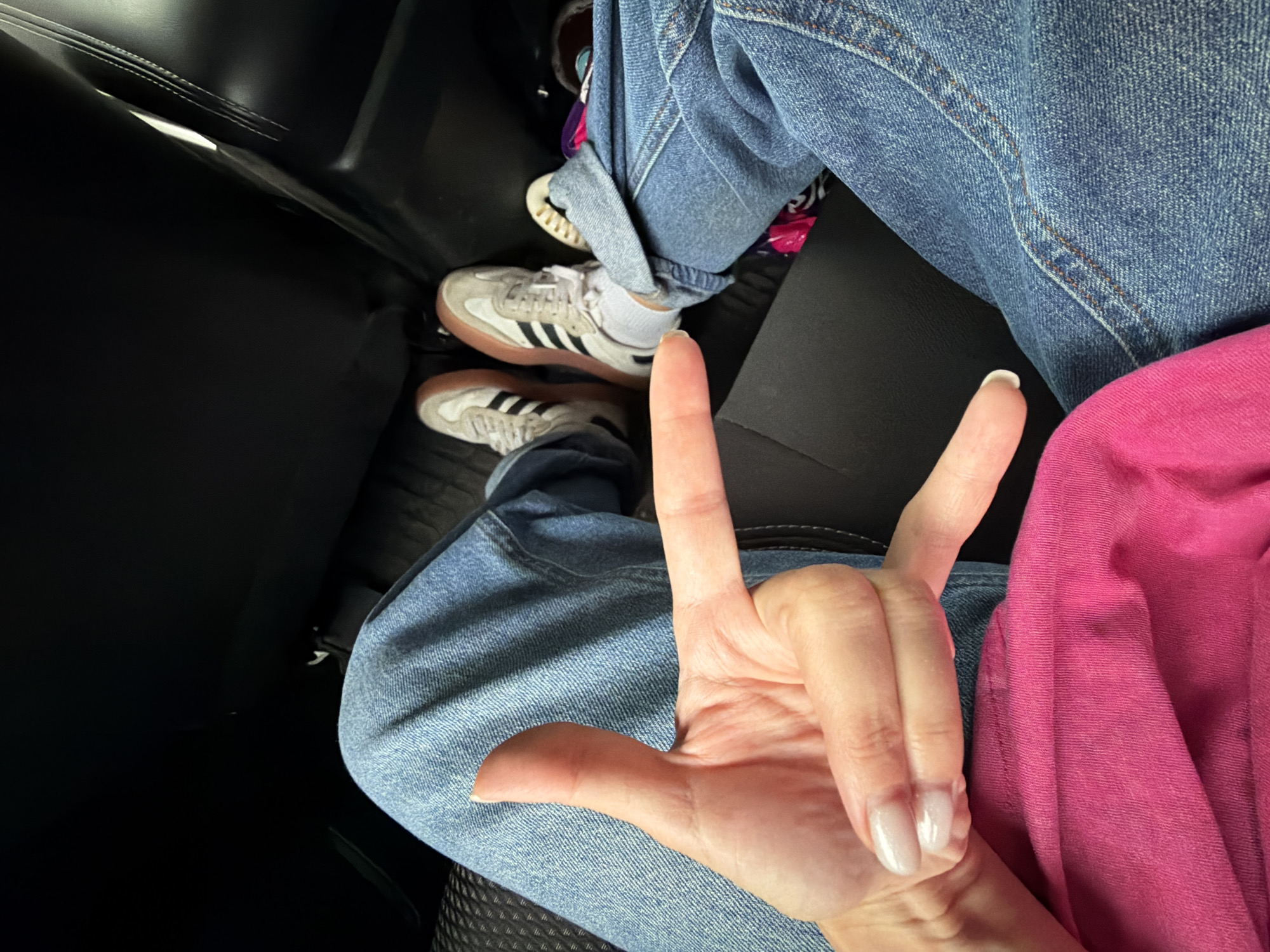 In a dimly lit vehicle interior, the image captures a close-up view of a person's hands and legs, suggesting they are seated. The person is wearing blue denim jeans and white sneakers with gum soles and black stripes. A bright pink garment, possibly a shirt or jacket, is visible in the foreground. The hand positioned in the center of the frame makes a "rock on" hand gesture, with the thumb and index finger extended and the other fingers curled. The background consists of dark upholstery, likely car seats. The scene's low lighting and the specific details of the clothing and hand gesture suggest an informal, candid moment. The image does not contain any discernible elements that would place it in Kimovsk, Russia.