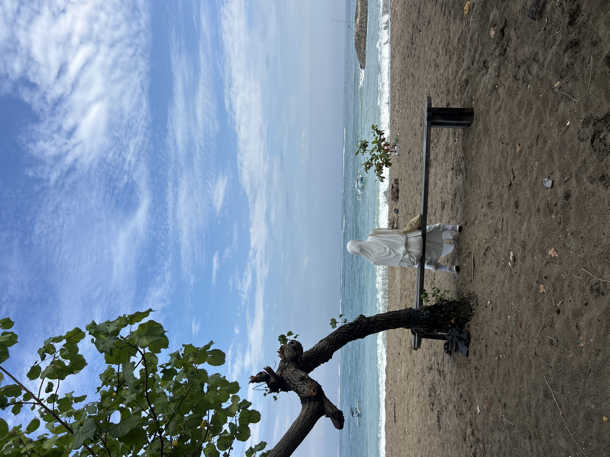 A person wearing a white hijab and white clothing sits on a wooden bench on a sandy beach in Kuta, Indonesia. The person is facing the ocean, which has gentle waves and is a turquoise color. In the distance, two small boats are visible on the water, and a stone jetty extends into the sea on the right side of the frame. A gnarled tree with green leaves is on the left side of the image, and a small green bush is to the right of the bench. The sky is blue with scattered white clouds. The sand on the beach is dark brown and appears wet in places. A straw bag is on the bench next to the person, and a pair of black sandals are on the sand near the base of the tree.
