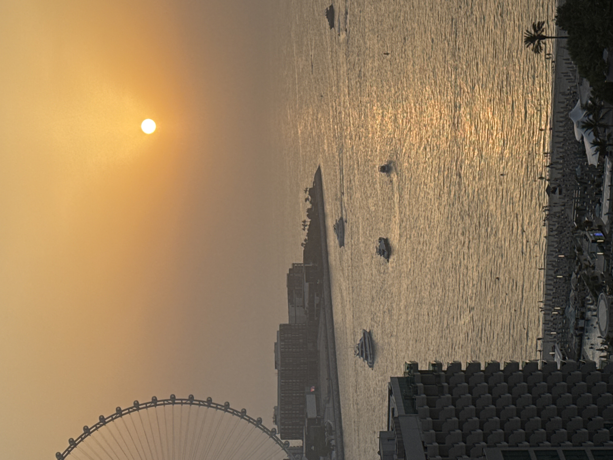 This image captures a coastal scene in Dubai, United Arab Emirates, likely during sunset. On the left, a portion of the Ain Dubai observation wheel is visible against a hazy, orange sky. In the middle ground, several boats are navigating the calm water. Further inland, a strip of beach is lined with numerous beach umbrellas and lounge chairs, with a scattering of people visible. To the left of the beach, a resort complex with swimming pools and a building with balconies is depicted. The overall atmosphere is one of a serene evening by the sea.