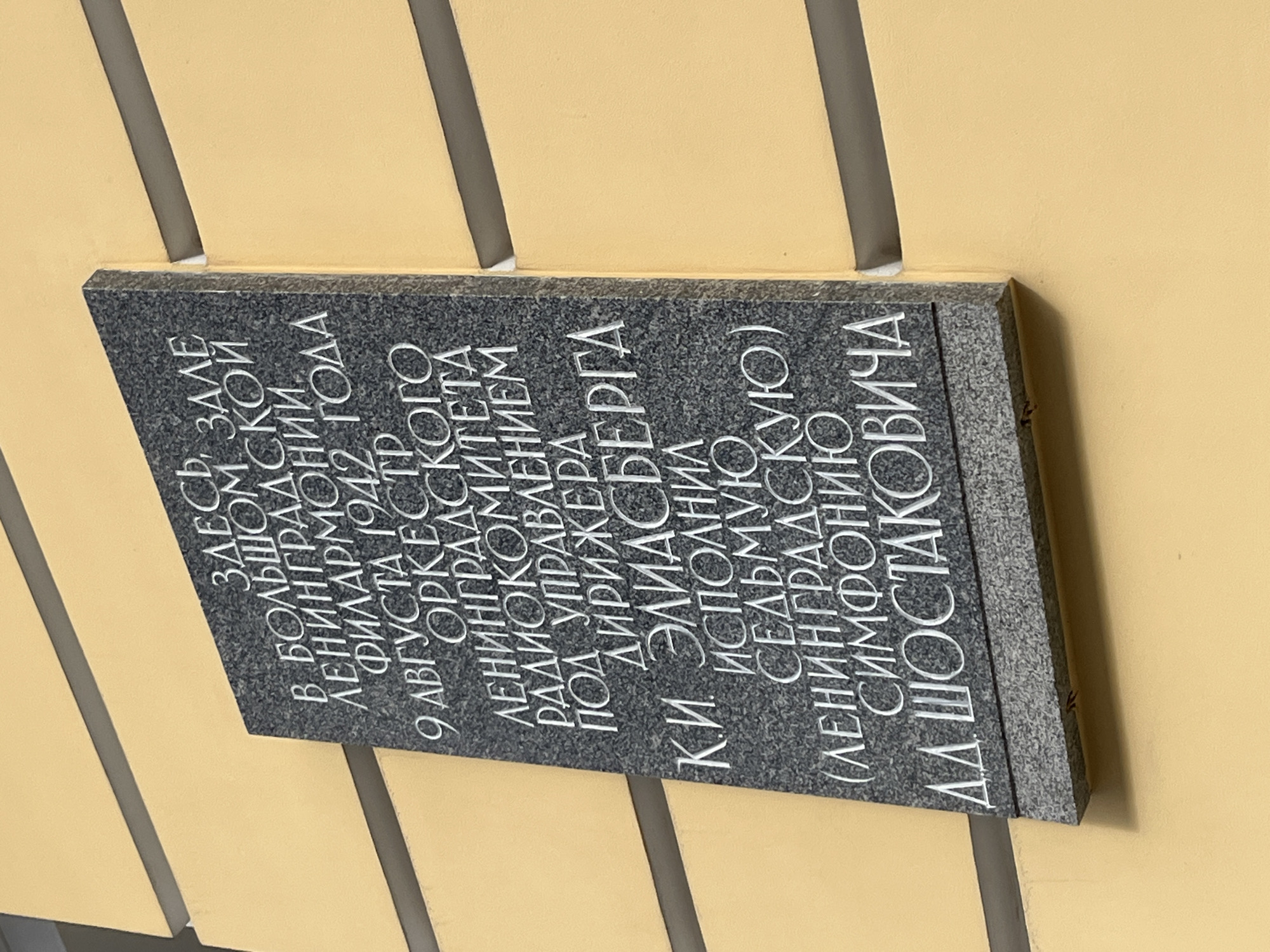 The image shows a gray stone plaque attached to a peach-colored wall. The plaque is engraved with white Cyrillic text. The text indicates that in this hall of the Leningrad Philharmonic, on August 9, 1942, the Leningrad Radio Committee Orchestra, under the direction of K.I. Eliasberg, performed the Seventh (Leningrad) Symphony by D.D. Shostakovich. The plaque is likely located in the city of Saint Petersburg, Russia, formerly known as Leningrad.