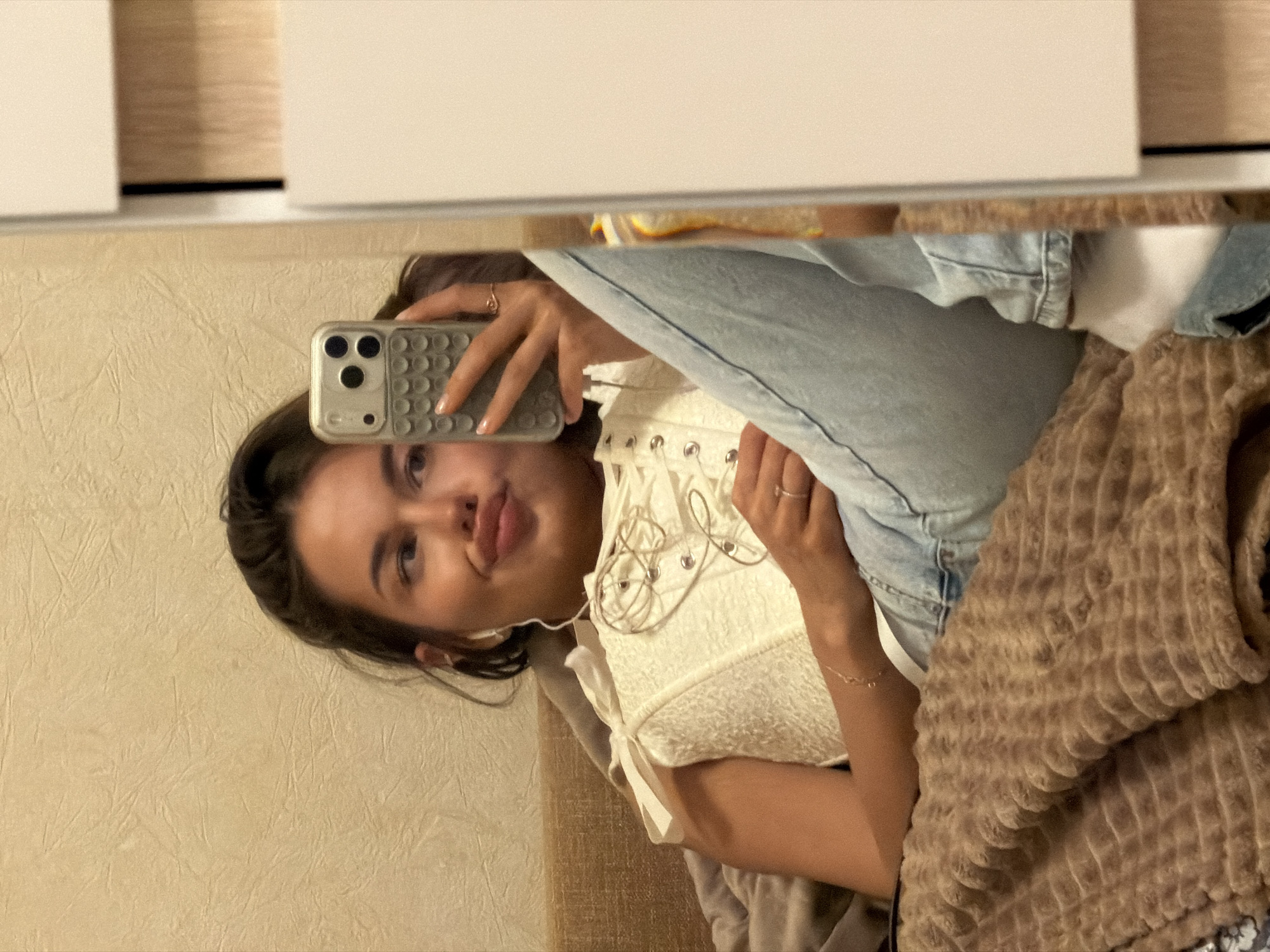 This media file captures a mirror selfie of a young woman taken indoors, likely in a bedroom or living area. The image is rotated 90 degrees counter-clockwise, showing the woman's head on the left and her body extending towards the right.
The central subject is a young woman, visible from the chest upwards. She is holding a light-colored smartphone, possibly silver or light gray, with a textured case and a triple-lens camera array, in her right hand to take the photo. She is looking directly at the phone's camera, making a playful pouting or "duck face" expression, with her head slightly tilted. She has dark hair, and her fingernails are painted with a light polish.
The woman is dressed in a light-colored, possibly cream or white, corset-style top featuring lacing details on the front. Light blue denim jeans are also visible, partially covering her lower body. She wears a simple ring on her right ring finger and a delicate chain bracelet on her right wrist.
The reflected background includes a textured, light beige or cream wall behind her head and shoulder. To her right, a soft, brown blanket or throw with a distinctive raised, ribbed texture is visible, suggesting she is reclining on a bed or couch. Above her in the reflection, a plain white surface, possibly a ceiling or upper wall, is seen with a thin brown horizontal line beneath it, which could be a shelf or frame. The lighting in the image suggests an indoor setting with ambient artificial light. There is no visible text in the image. While the location context is given as Moscow, Russia, this is not discernible from the visual content of the image.