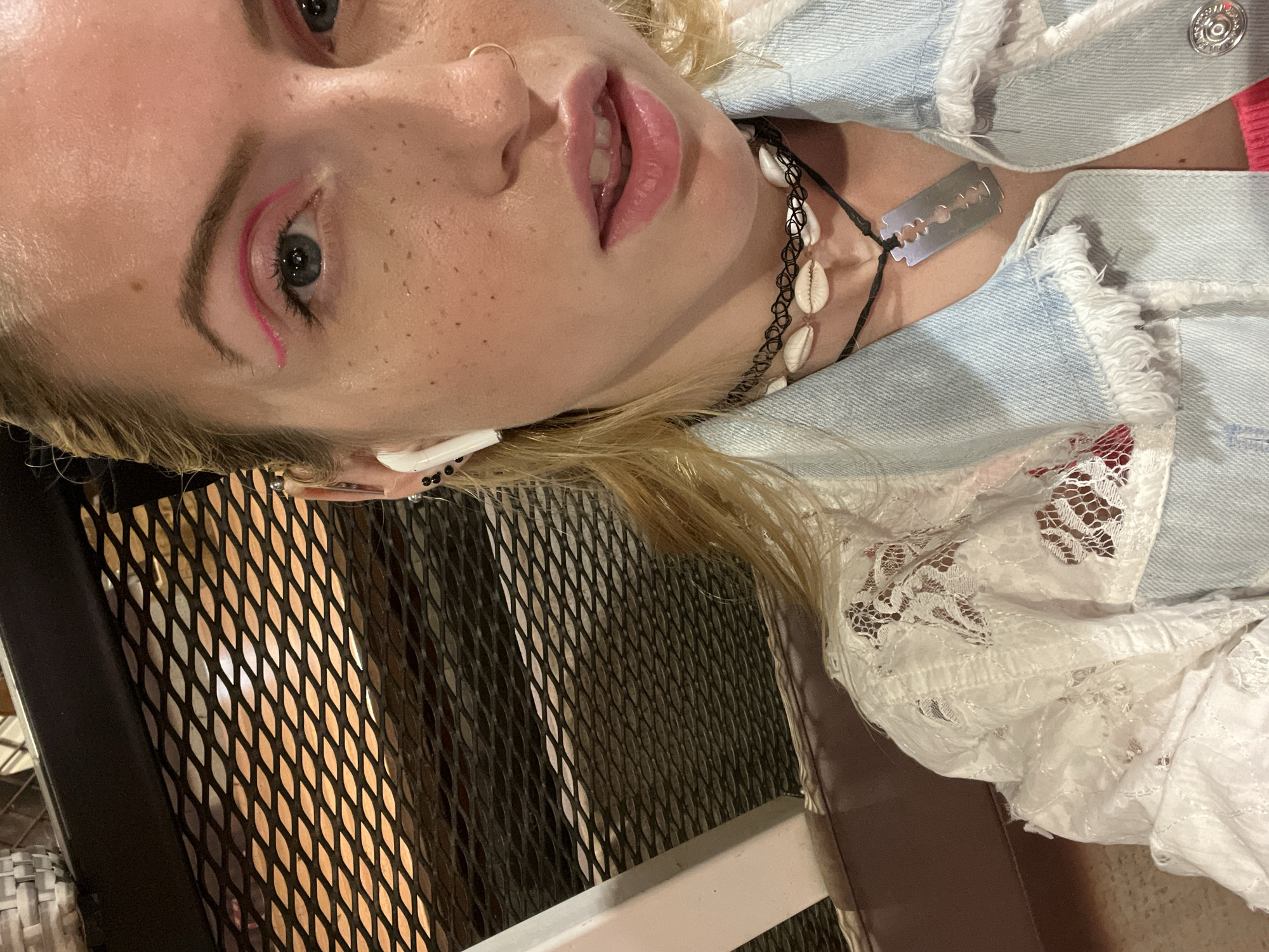 A young woman is shown in a close-up, partially rotated view. She is fair-skinned with freckles, blonde hair, and light blue eyes with pink eyeliner. She wears an in-ear earbud in her left ear, a nose ring, and two necklaces: one is a black choker with seashells, and the other appears to have a razor blade pendant. She is wearing a light blue denim jacket with white lace detailing at the sleeve and a pink top underneath. The background includes a lattice metal structure and the edge of what seems to be a seat. No information in the image indicates that the location is Plovdiv, Bulgaria.