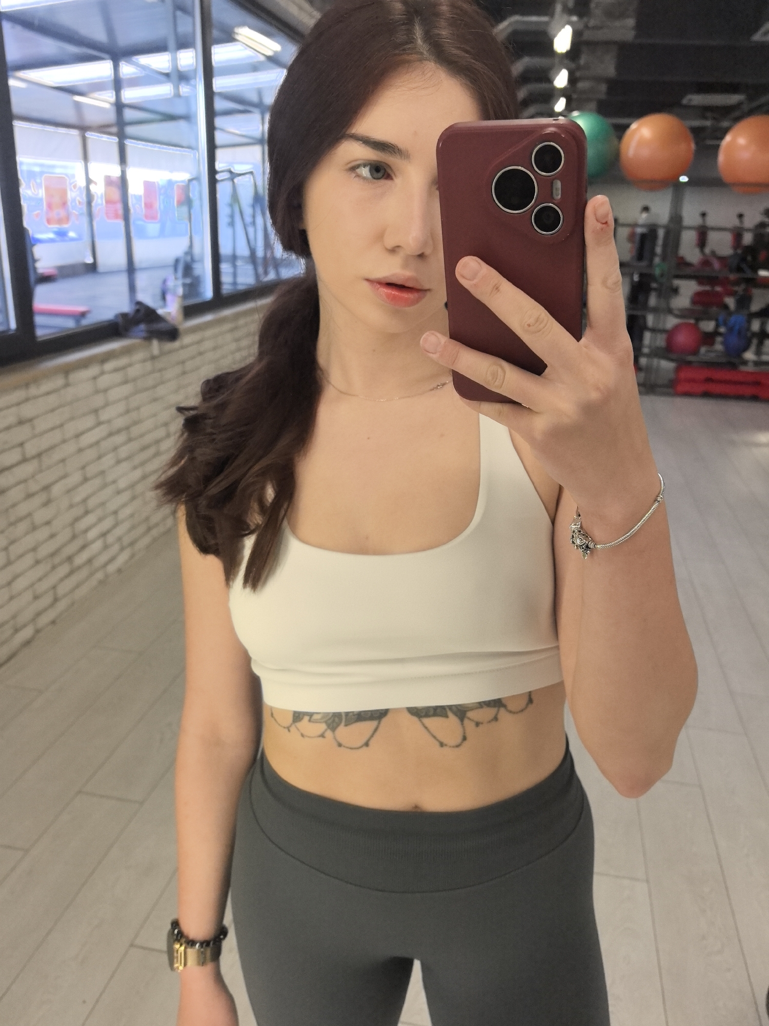 A young woman with brown hair is taking a selfie in what appears to be a gym, identified by exercise equipment in the background. She is wearing a white sports bra and gray leggings. She has a watch on her left wrist and a bracelet on her right. A tattoo is visible just above the waistband of her leggings. The background includes exercise equipment and exercise balls. The gym appears to be located in an urban area, with views of buildings visible through the windows. While Stara Zagora, Bulgaria, is not directly visible, it is assumed to be the location based on other information.