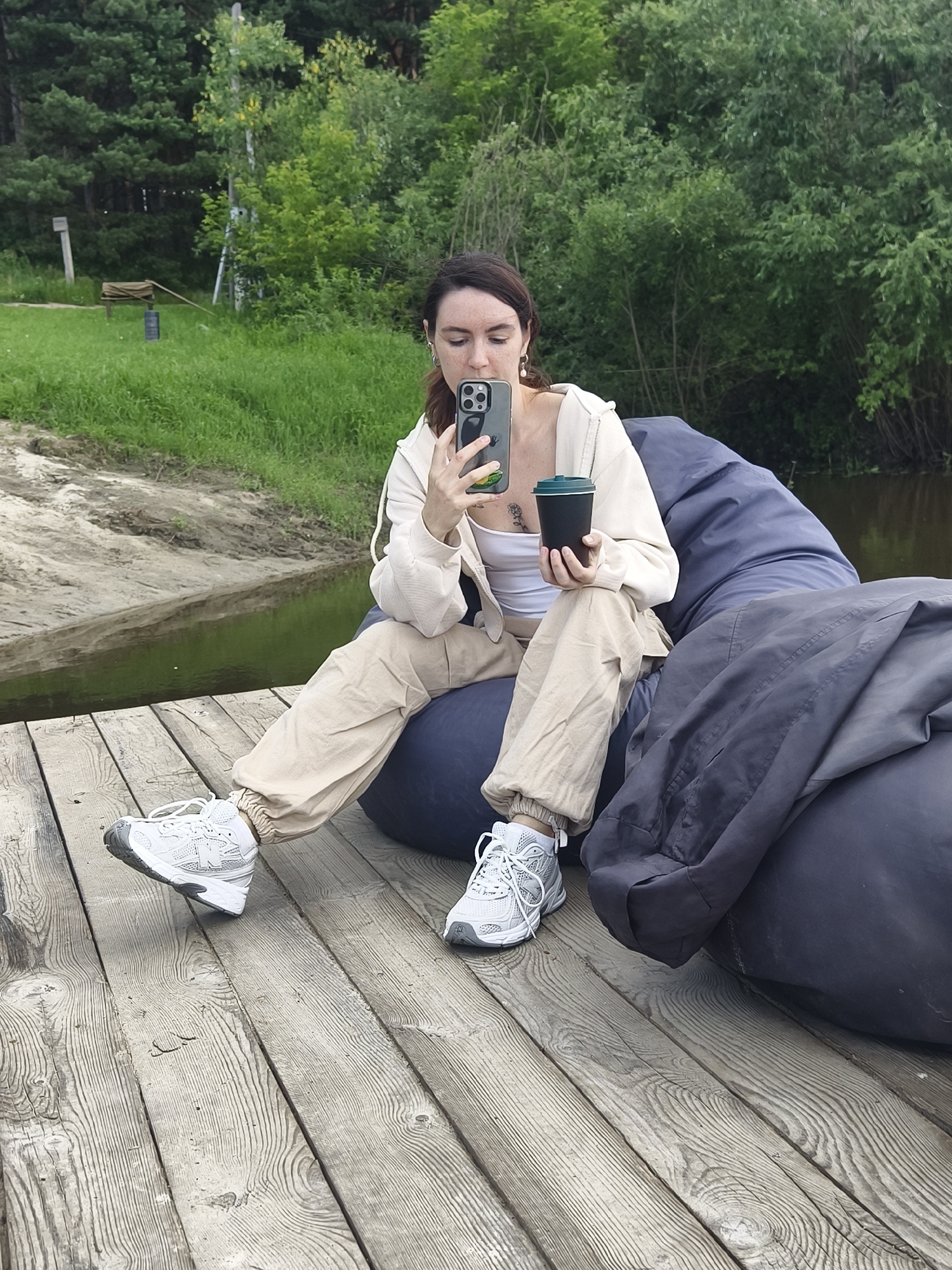 In Онохино, Russia, a young woman with dark hair is seated on a large, dark blue beanbag chair on a wooden pier. She is wearing a white tank top, a light beige hoodie, and loose beige cargo pants. Her feet are clad in white and grey athletic shoes. In her left hand, she holds a black disposable coffee cup, and in her right hand, she is holding a smartphone, appearing to take a selfie or photograph. The pier extends over a body of calm water, with a muddy bank on the left and dense green foliage and trees forming the background. The weather appears to be overcast, with diffused natural light.