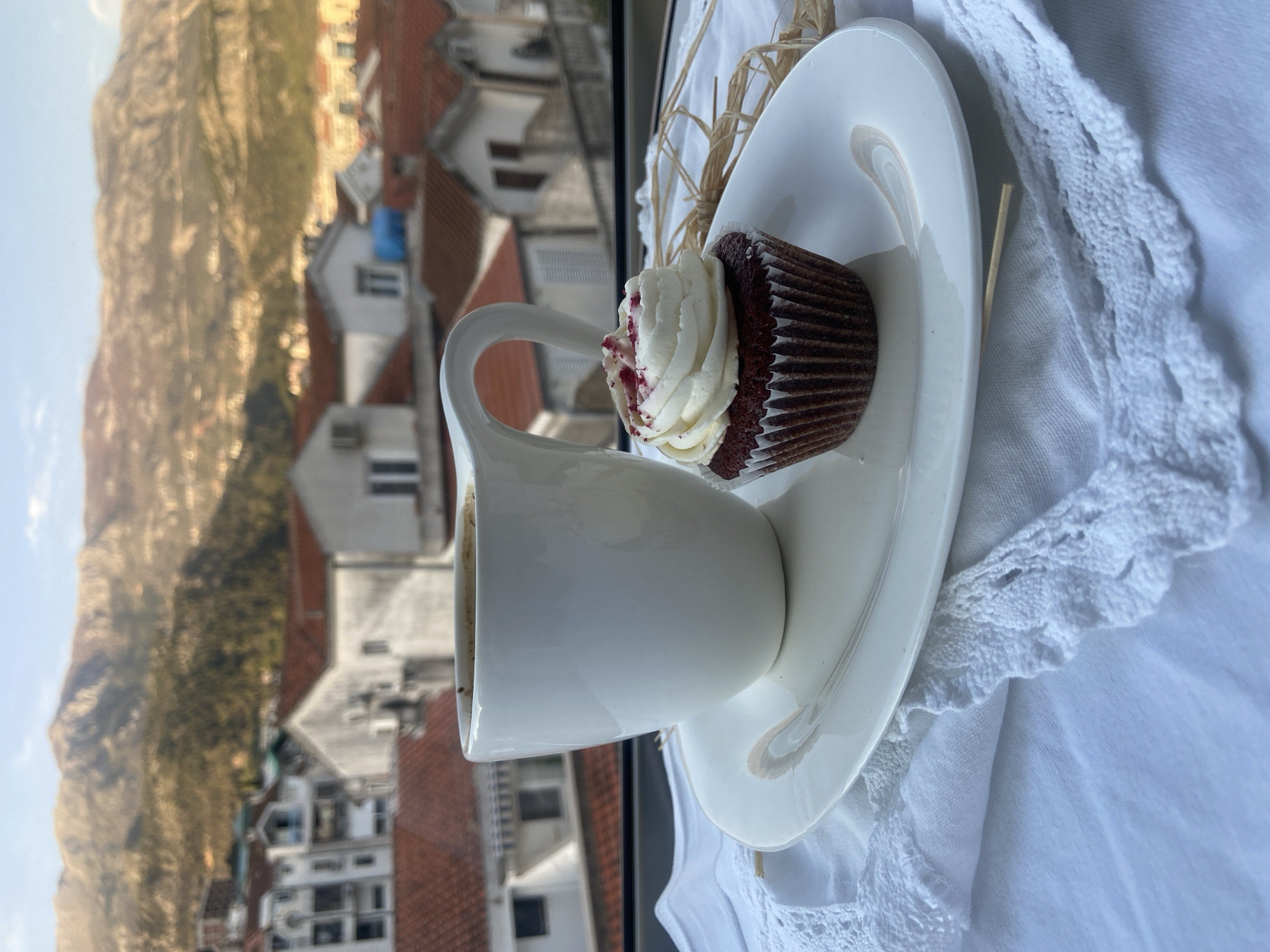 A slightly rotated, high-angle, eye-level shot depicts a white coffee mug on a matching saucer and a cupcake on a white crocheted tablecloth, with a cityscape visible through a window.
The white coffee mug, filled with dark liquid, sits on the left side of the saucer with its handle facing towards the camera. A chocolate cupcake with white frosting, speckled with red crumbs, rests on the right side of the saucer. The saucer is placed on a white crocheted tablecloth, which adds texture to the foreground.
In the background, through the window, is a view of Budva, Montenegro. The architecture consists of closely packed buildings with red tile roofs. In the distance, a mountain range rises from the city, and the sky is a pale blue. The overall scene suggests a relaxing coffee break with a scenic view.