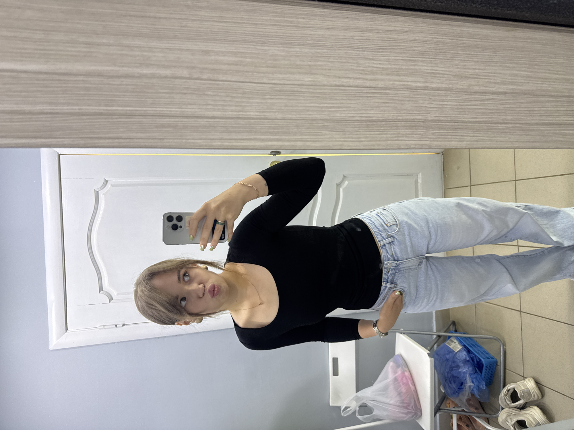 A young woman with light brown hair and bangs stands in what appears to be a room with pale blue walls and a wooden-paneled door in the background. She is wearing a black long-sleeved shirt and light-wash denim jeans. She is taking a mirror selfie with a smartphone held in her right hand, and her left hand is tucked into her front jeans pocket. Her facial expression is one of playful puckering. She has a delicate gold necklace on, and a bracelet on her left wrist. To her left, there is a white folding chair with a plastic bag containing items on it, and a blue basket with more plastic-wrapped items below it. A pair of white sneakers is visible on the floor in the foreground. The scene has a casual, domestic feel. This image was taken in the city of Chaikovsky, Russia.
