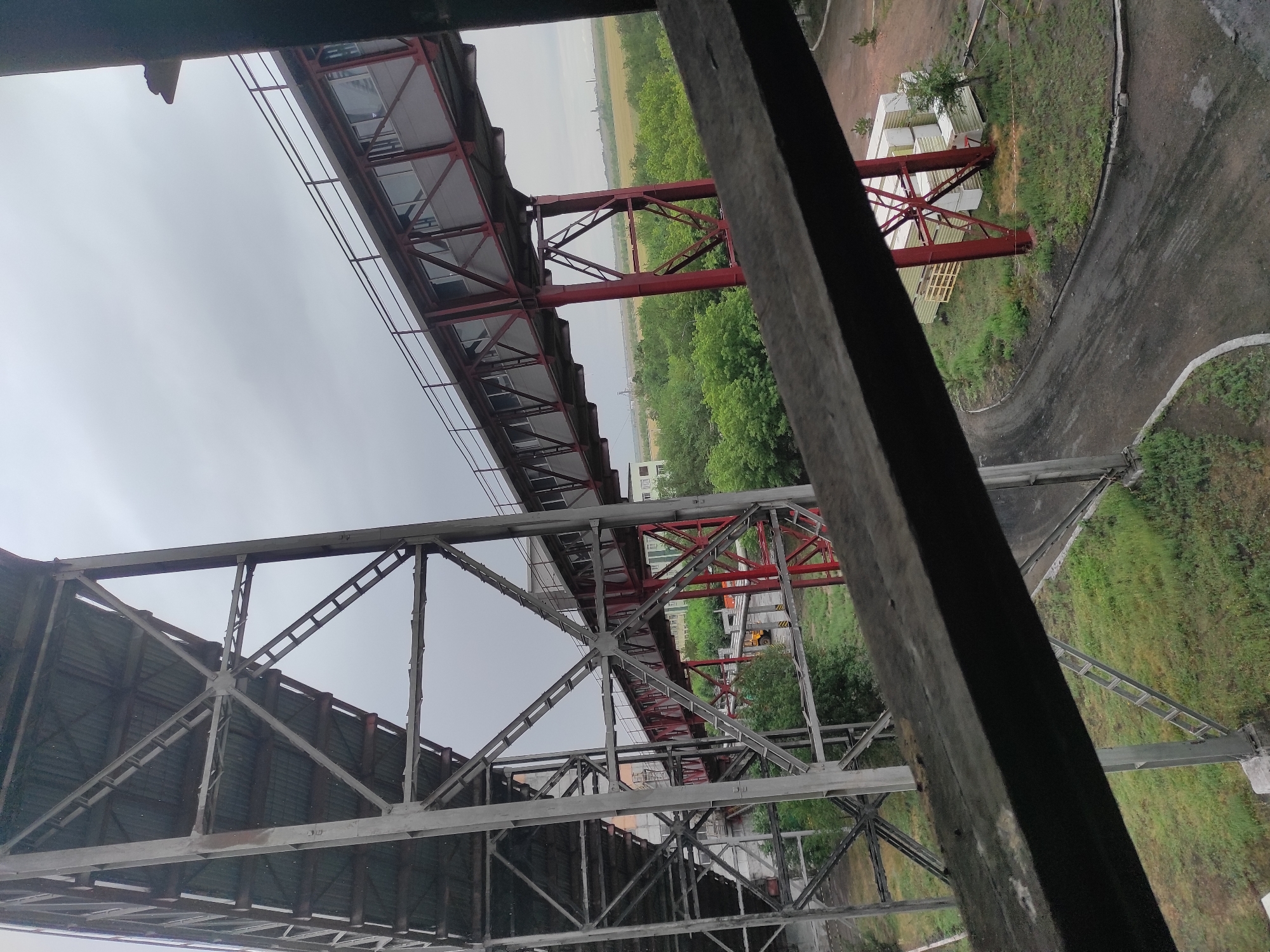 The image captures an industrial scene dominated by a large, gray metal structure on the left, likely part of an older industrial conveyor system. Extending from this structure and across the frame is a red and gray elevated walkway or conveyor. This walkway has large windows along its length and is supported by red metal beams. The ground below the walkway is covered in green grass and a dirt road. In the background, a line of trees separates the industrial area from a flat, open landscape under a cloudy sky. Some building materials, possibly pre-fabricated structures, are stacked to the right of the red support beams. The overall impression is one of an active or recently active industrial site. The location is identified as Kyzyl, Kazakhstan.