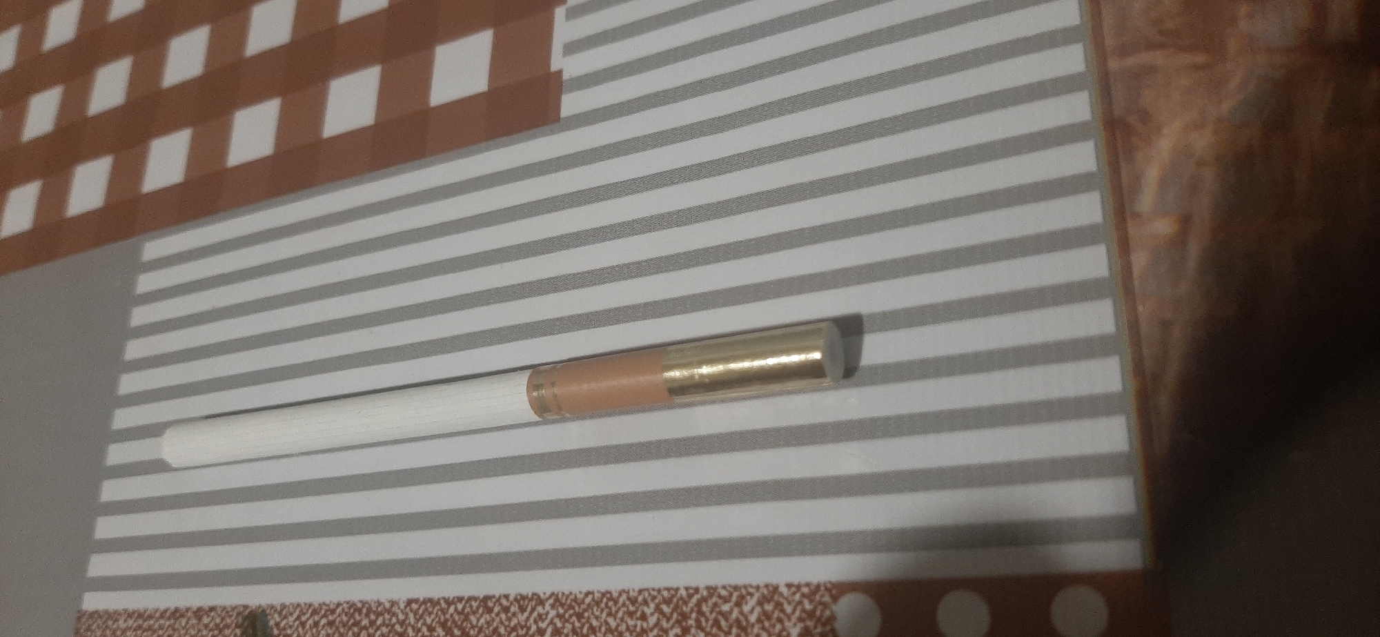 A single cigarette lies on a striped surface. The cigarette has a white filter end and a tan filter end, with a golden band separating the two. The surface is a light gray with thin, horizontal dark gray stripes. In the upper left corner of the image, a section of a brown and white checkered pattern is visible. The background to the right of the striped surface is a blurry, dark brown textured surface. There are no people or actions depicted in the image. The location in the city of Yessentukskaya, Russia, is not discernible from the image.