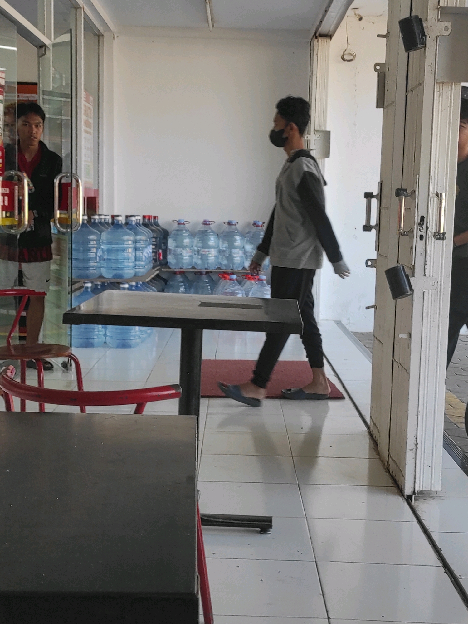 In an outdoor, covered walkway in Purwodadi, Indonesia, a young man wearing a black face mask, a grey and black long-sleeved shirt, and black pants walks towards the right side of the frame. He is wearing blue slip-on shoes. In the background, a rack of blue-capped water jugs is visible. To the left, another young man in a red shirt and dark jacket stands near a glass door. A red chair is visible in the foreground. A dark-colored table is positioned in the middle ground. The floor is tiled.
