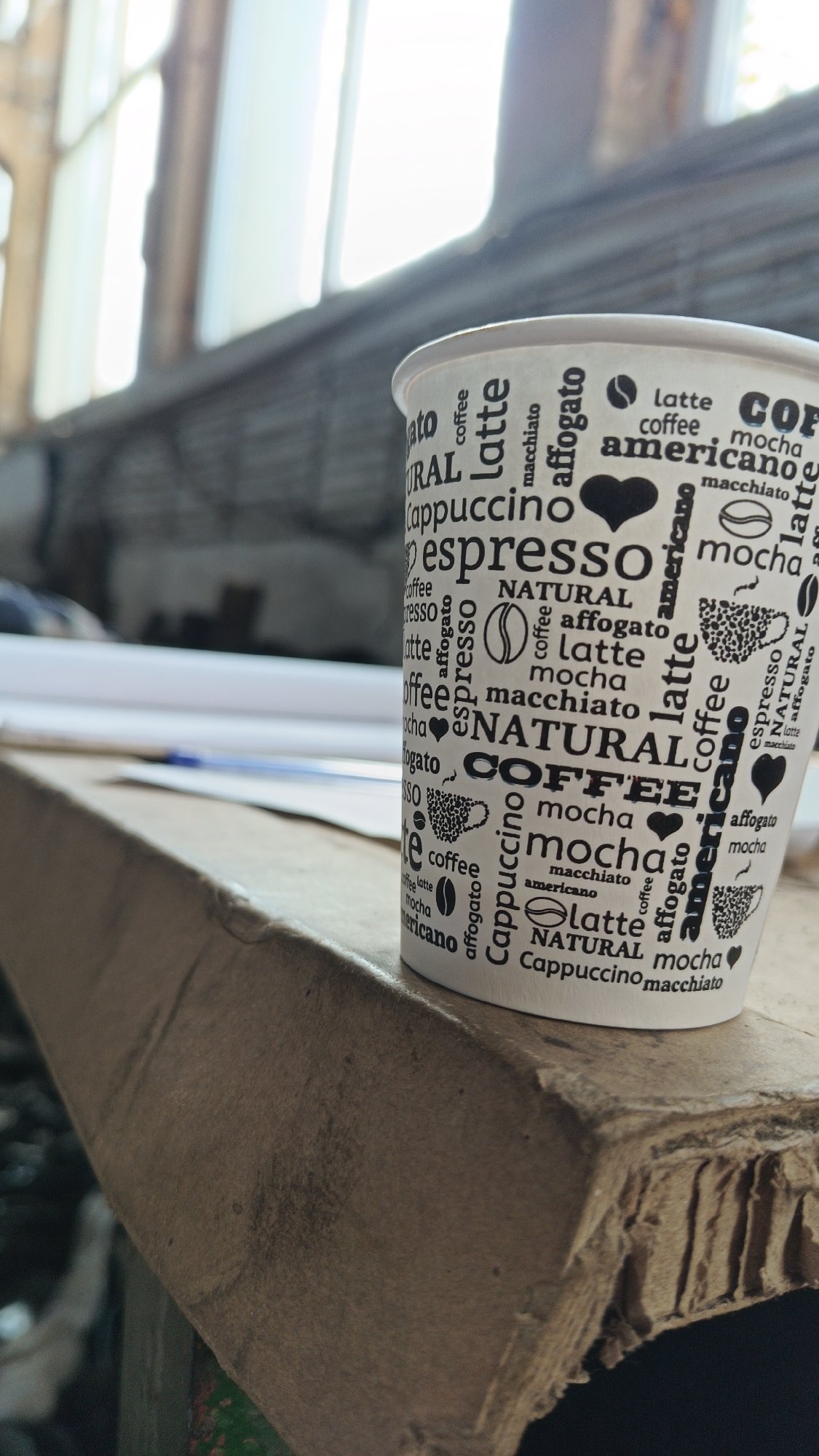 A close-up shot shows a white paper coffee cup, printed with various coffee-related words and illustrations, resting on the edge of a corrugated cardboard surface. A blue pen and some papers are partially visible in the blurred background. The scene is set near a window, evidenced by the diffused light and visible window frame. The location appears to be a cluttered or possibly a workspace.