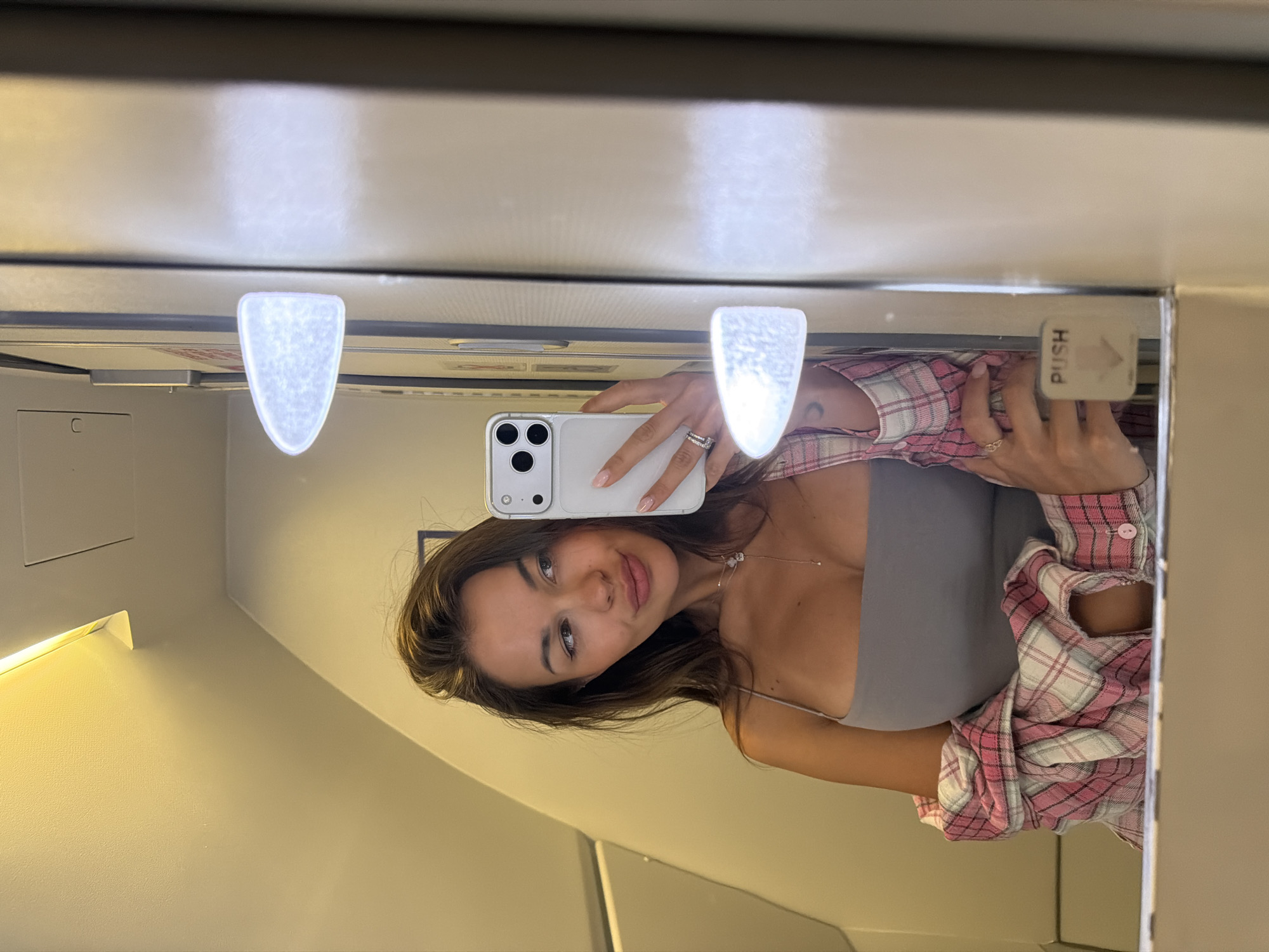 A woman is taking a selfie in a mirror. She is holding a white iPhone. She is wearing a grey top and a pink and white plaid shirt. The mirror appears to be in a bathroom, possibly on an airplane. Above the mirror are two small lights and a "PUSH" sign. The location is unknown and cannot be determined from the image. The city of Krasnoyarsk, Russia, is not indicated in any way.