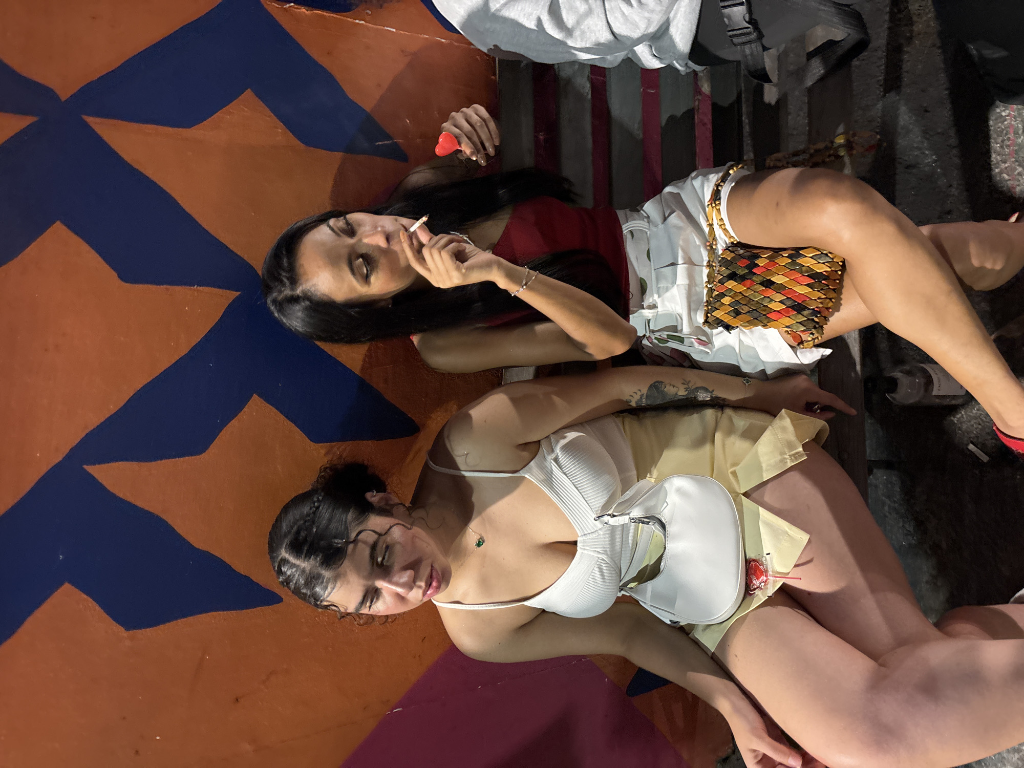 Two women are lying on a painted surface of orange, blue, and magenta in what appears to be a public space. One woman has long, dark hair and wears a red top and white shorts. She is smoking a cigarette and holding a red heart-shaped object. A colorful purse rests on her leg. The other woman has dark hair styled in a bun and wears a white top and yellow shorts. She carries a white purse. Both women appear relaxed and are looking at the camera. The photo is taken outdoors at night. Based on the image content, it is impossible to determine the specific location in São Paulo, Brazil.