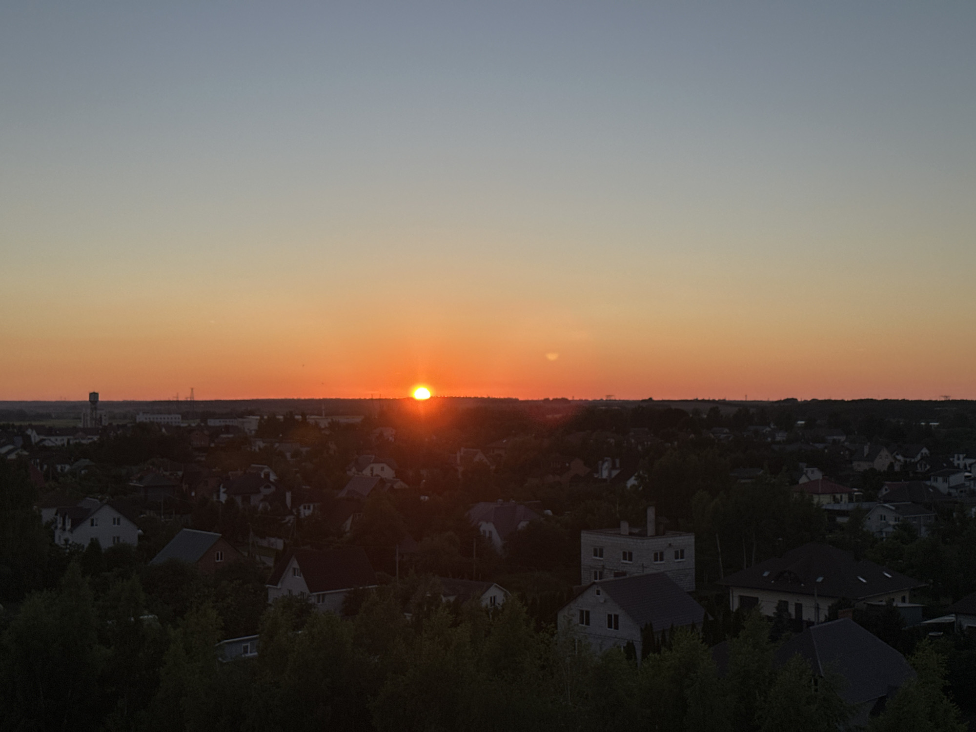 The image captures a sunset over a residential area in Shchomyslitski Sel'skiy Sovet, Belarus. The sun, a bright orange orb, is low on the horizon, casting a warm glow across the sky which transitions from a pale blue at the top to a vibrant orange near the horizon. Below the sky, a dense collection of houses with various roof shapes and colors are visible, interspersed with numerous trees that cast dark shadows. In the distance, a tall water tower stands out against the setting sun, along with some industrial or utility structures. The overall scene depicts a tranquil evening in a suburban or rural landscape.