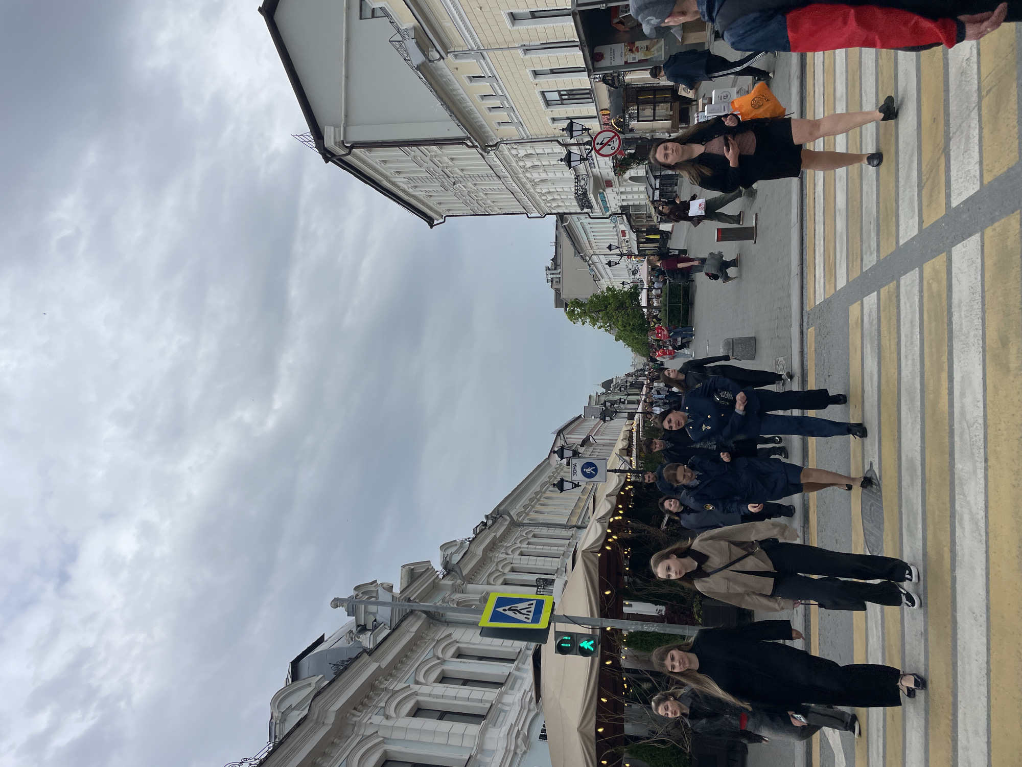 The image depicts a street scene in Kazan, Russia. A pedestrian crosswalk is visible with people crossing the street. Buildings line both sides of the street. A green pedestrian crossing signal is illuminated. The sky is overcast. People of various ages and wearing different clothes are present.