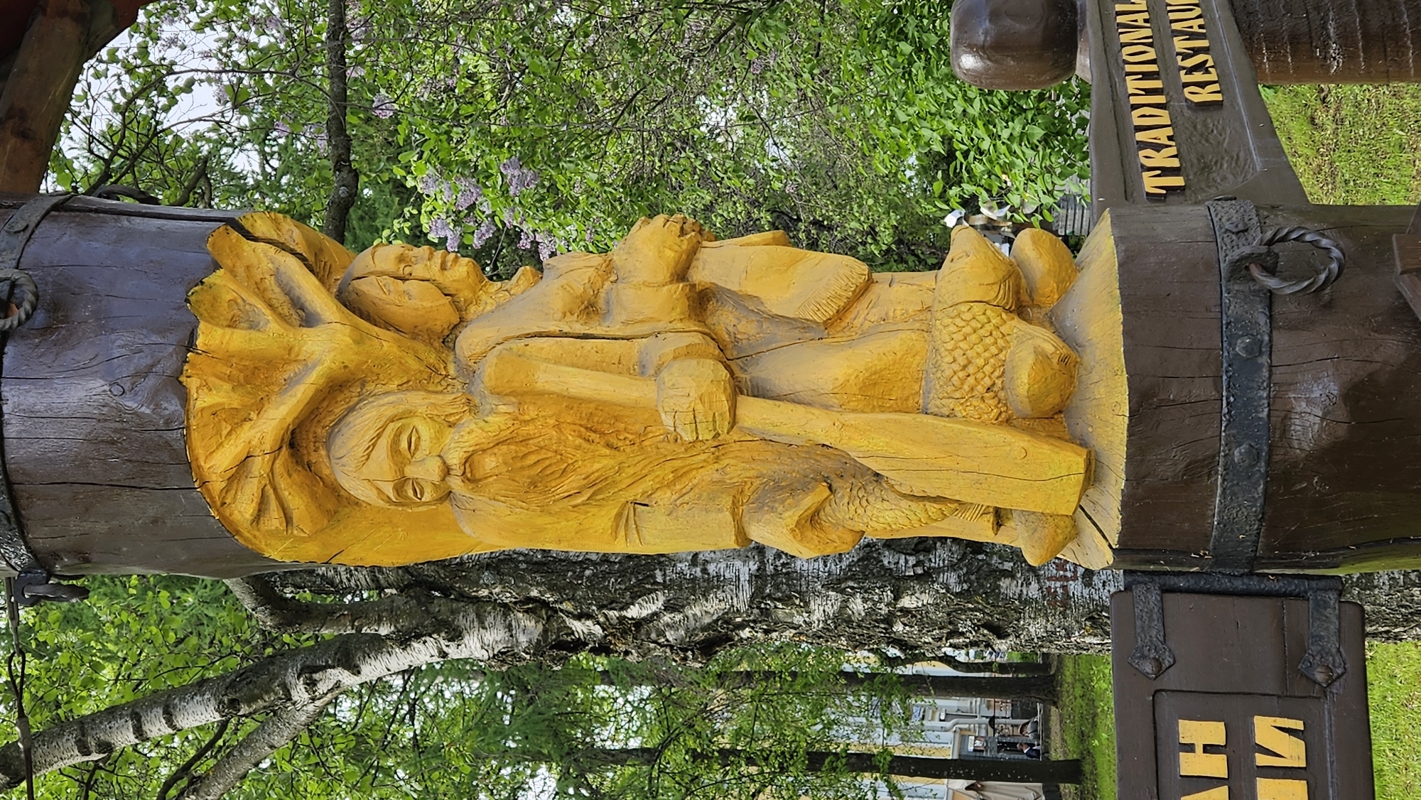 A wooden carving of two people, likely depicting a scene from a traditional story, is affixed to a wooden barrel that functions as part of a sign for a "Traditional Restaurant." The carving is painted yellow, while the barrel is brown with black metal straps. The sign is located in a wooded area, with trees visible in the background and around the structure. The exact location is in the city of Unknown, Unknown.