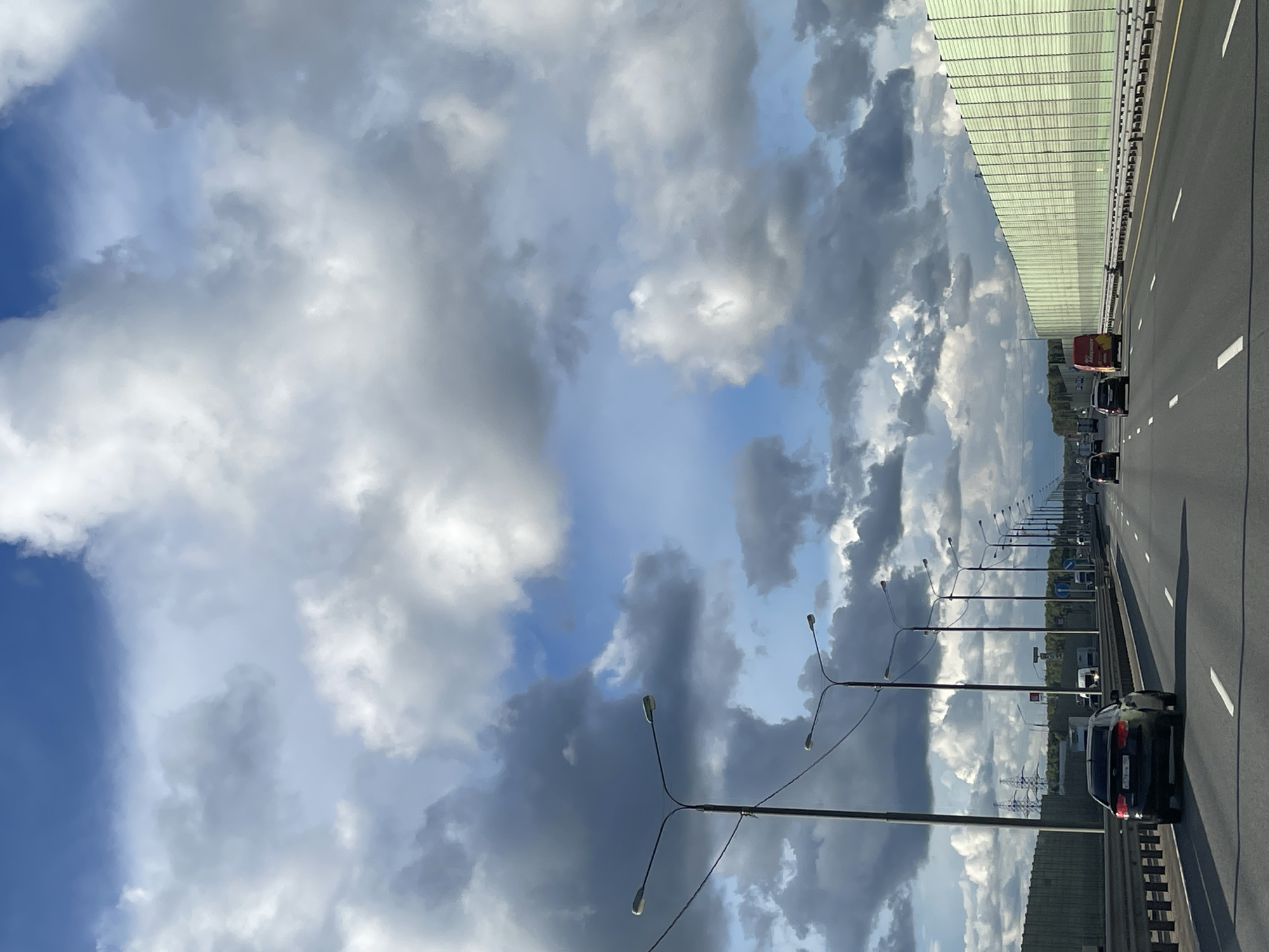The image shows a view from a bridge or overpass, oriented vertically, with a highway stretching into the distance. Several vehicles are visible on the roadway, including a red and white van, a black sedan, and other cars further away. Streetlights line the side of the highway. The sky is filled with a mix of white and dark clouds with patches of blue visible. The location is potentially in Berezhki, Russia. A sound barrier runs along the side of the highway in some sections.