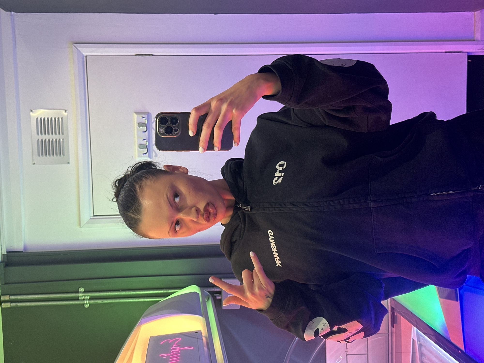 A female individual is depicted taking a selfie inside a tanning booth. She has dark hair tied in a bun, pursed lips, and is looking towards her left. She wears a black zipped hoodie with "GYMSHARK" (reversed) printed in white on the chest and a white circular logo featuring a masked face (associated with "SPO") on the left sleeve. Her right hand holds a dark-colored smartphone, likely an iPhone, capturing the image. Her left hand is raised, making a "rock on" gesture, with light-colored nail polish visible. A small tattoo is present on her left inner wrist. To the left, a tanning bed or vertical tanning unit is partially visible, emitting prominent pink, green, and blue neon lights. Mirrored pink text, likely "milyn" or part of a brand name, appears on the tanning unit's surface. The wall behind the tanning unit is dark green with two metallic conduits. To the right, a white door with white trim is present, featuring a silver rectangular ventilation grille mounted above it and three silver coat hooks. The environment is illuminated by ambient neon lighting, casting a strong purple hue on the right side of the scene. The location is a tanning booth within SPO – Fitness and Wellness, located at Unit 2, The Pantiles, Tunbridge Wells, TN2 5TE, United Kingdom.