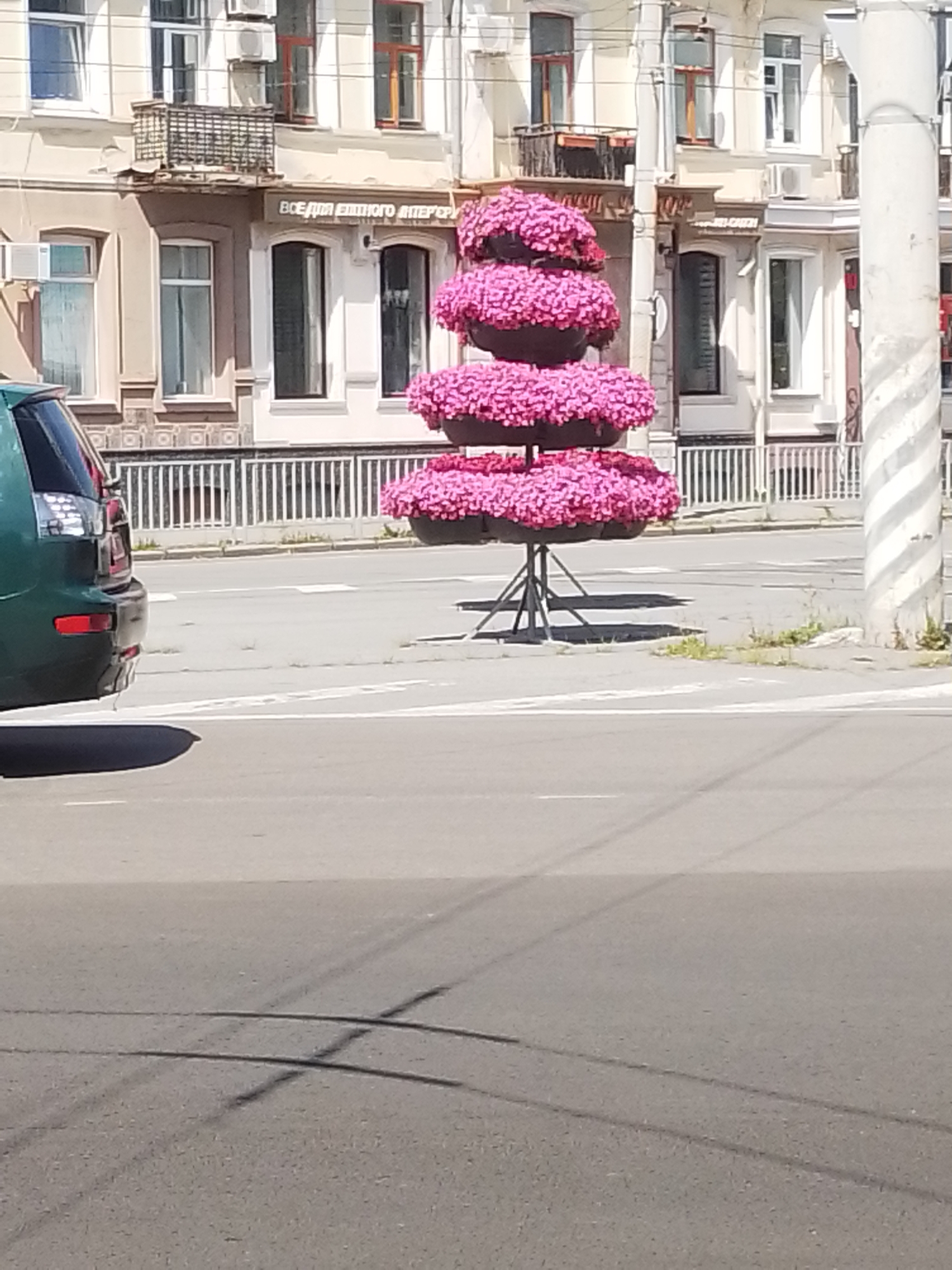 A three-tiered planter filled with pink flowers sits on a tripod stand on a sidewalk. The planter is decorated with pink flowers that cascade down its sides. Behind the planter, a building with several windows and a balcony is visible. A green car is parked on the side of the road. The scene appears to be in Poltava, Ukraine.