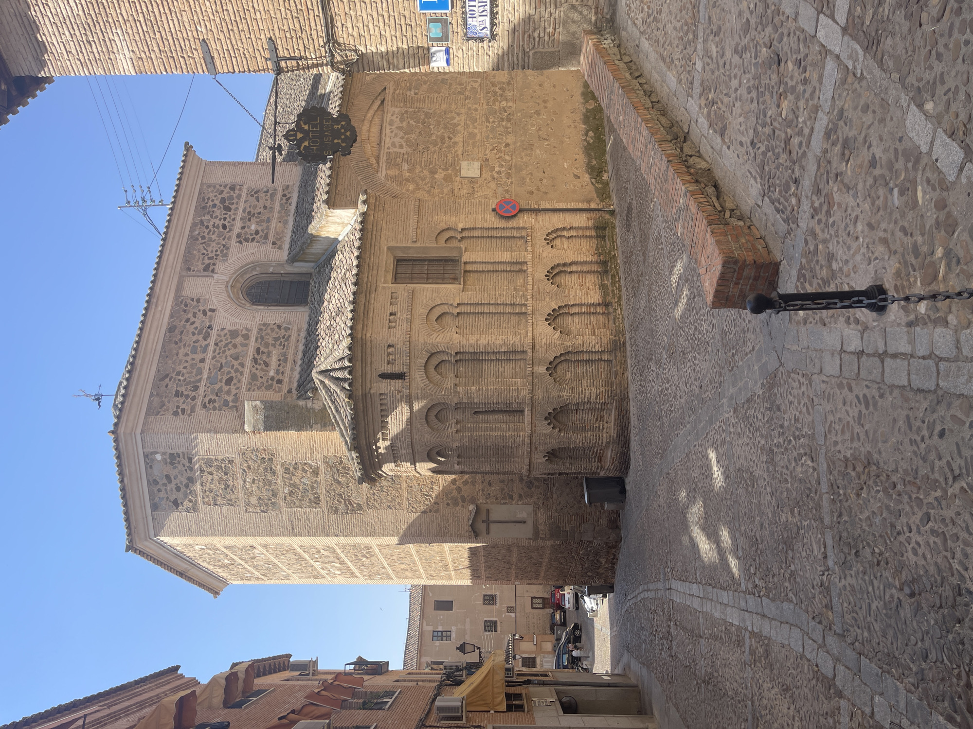 This is a ground-level, outdoor, eye-level shot of a historic street in Toledo, Spain, under a clear blue sky. The scene is dominated by an old, multi-sided building constructed of brick and stone, with a tiled roof. The building has arched window and decorative brickwork. A sign reading "HOTEL S ISABEL" hangs from a bracket on its side. Adjacent to this building is another brick structure, taller and more modern in appearance.
The street is paved with cobblestones and features a low, brick-lined planter. There are street signs, including one with a red circle and a diagonal line, indicating no parking. A dark metal bollard connected to a chain is visible in the foreground. A trash bin is placed near the base of the larger building, and a cross is affixed to its wall. In the background, several cars are parked on the street. The architecture suggests a medieval or early modern period, typical of Toledo's historic districts.
