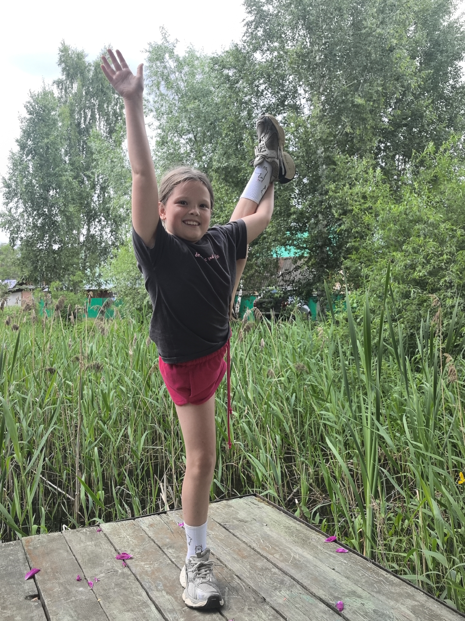 A young girl stands on a wooden platform, performing a vertical split with one leg extended straight up. She is wearing a dark t-shirt, pink shorts, white socks with a drawing of a cat, and athletic shoes. Her left arm is raised in the air, and she smiles at the camera with a joyful expression.
The setting is outdoors, surrounded by tall green reeds, suggesting a marshy or lakeside environment. In the background, there are trees and hints of buildings, possibly houses, with a green fence visible. The lighting indicates it is daytime, with overcast or diffused light, as there are no harsh shadows. The overall mood is cheerful and energetic, capturing a moment of athletic prowess and happiness. The location is identified as Noviy, Russia.