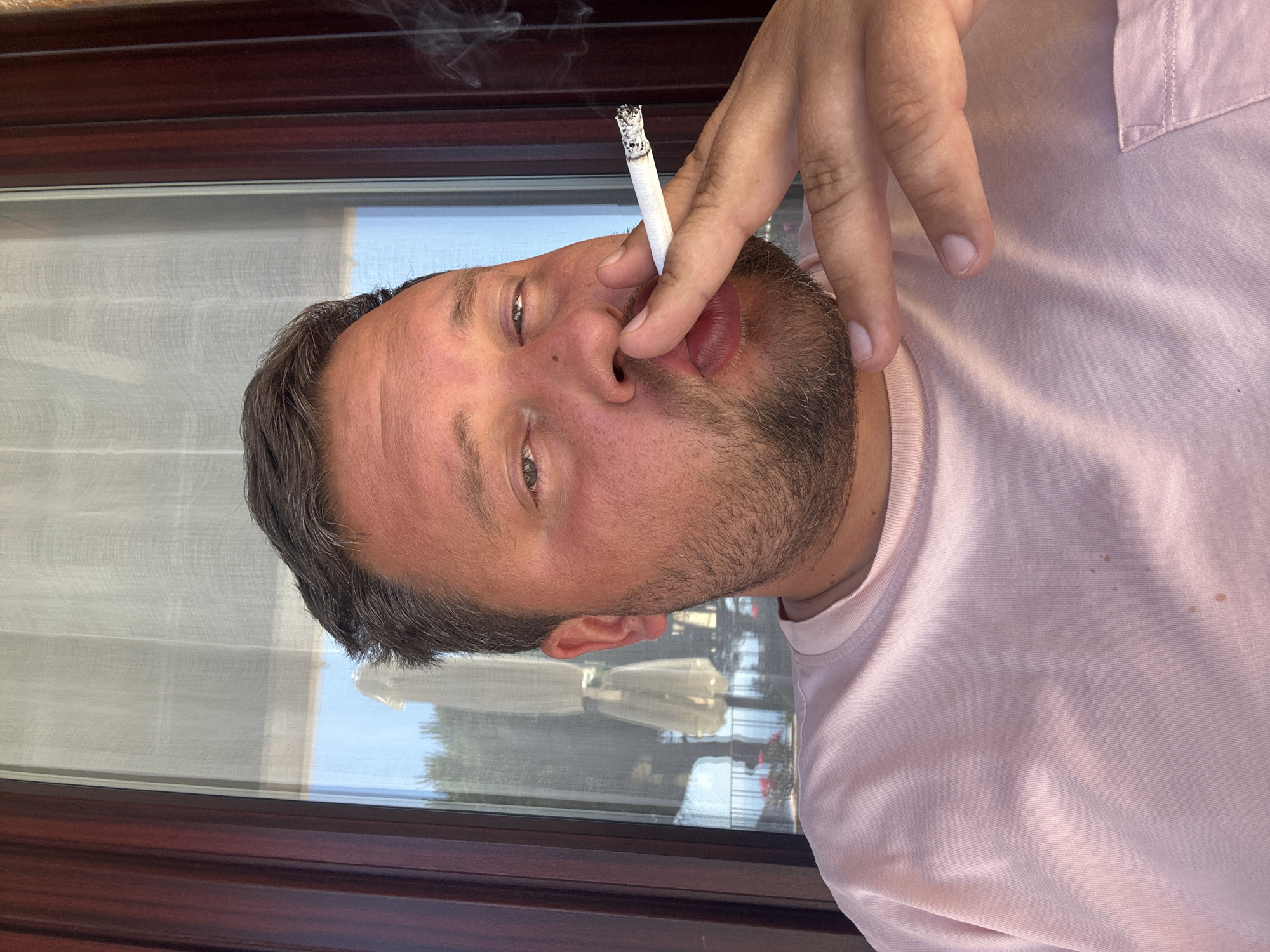 A man with short brown hair and a beard is holding a lit cigarette to his mouth. He is wearing a pink t-shirt. He appears to be sitting in a restaurant or bar with a view of the city of Sant Feliu de Guíxols, Spain, which is visible through a window. The reflection in the window shows outdoor seating and foliage.