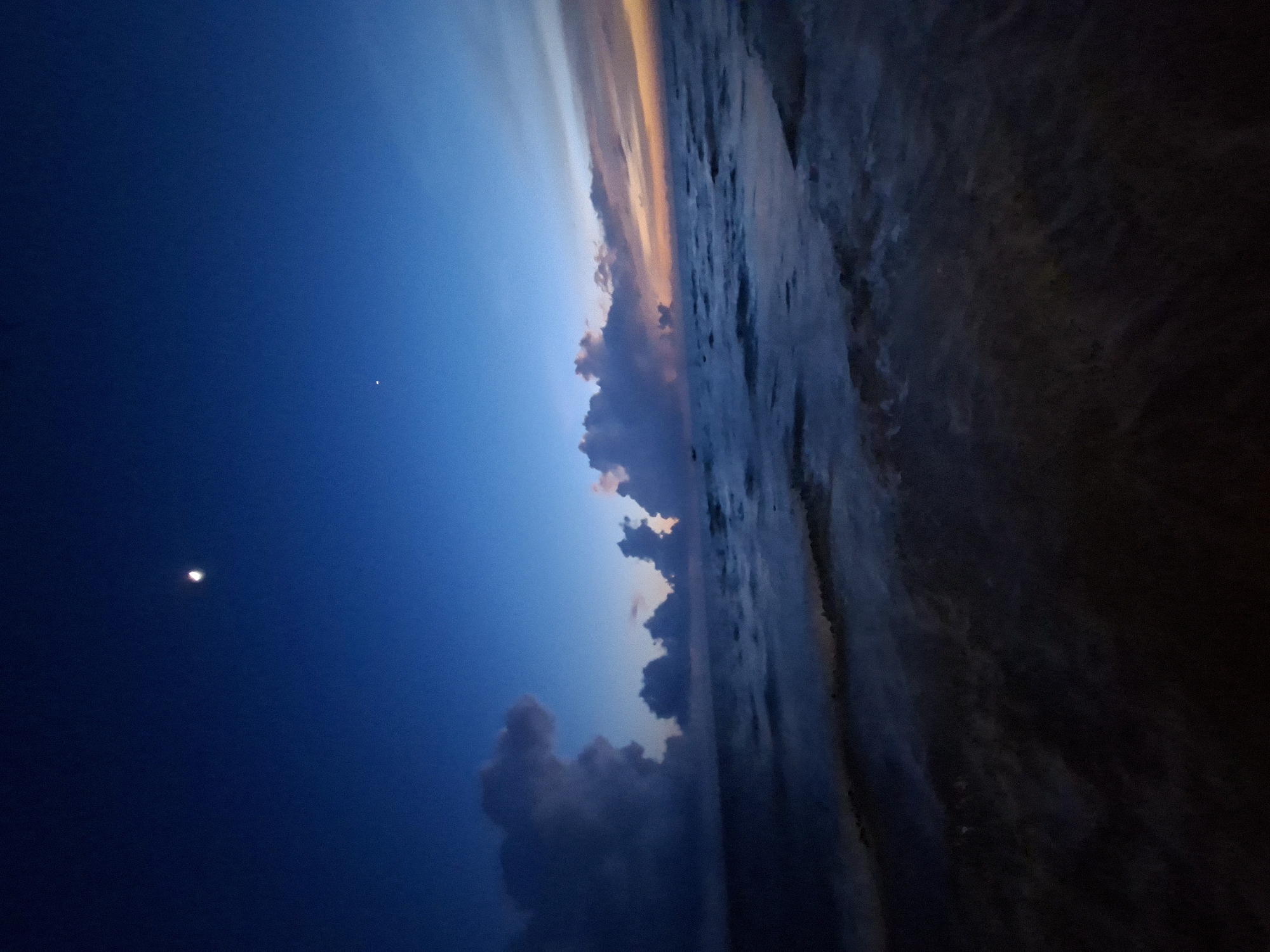 This is a wide-angle, low-light photograph of a seascape at twilight. The sky is a deep blue, transitioning to lighter shades of blue and hints of orange and pink near the horizon. A sliver of the moon is visible in the upper left quadrant of the sky, and a small, bright star is to the right of the moon. Dark, dramatic clouds are scattered across the sky, particularly on the left side. The sea is dark with visible waves, and the water reflects the colors of the sky. The foreground is a dark, indistinct shoreline or shallow water. There are no people or identifiable objects in the scene. The location is unknown.
