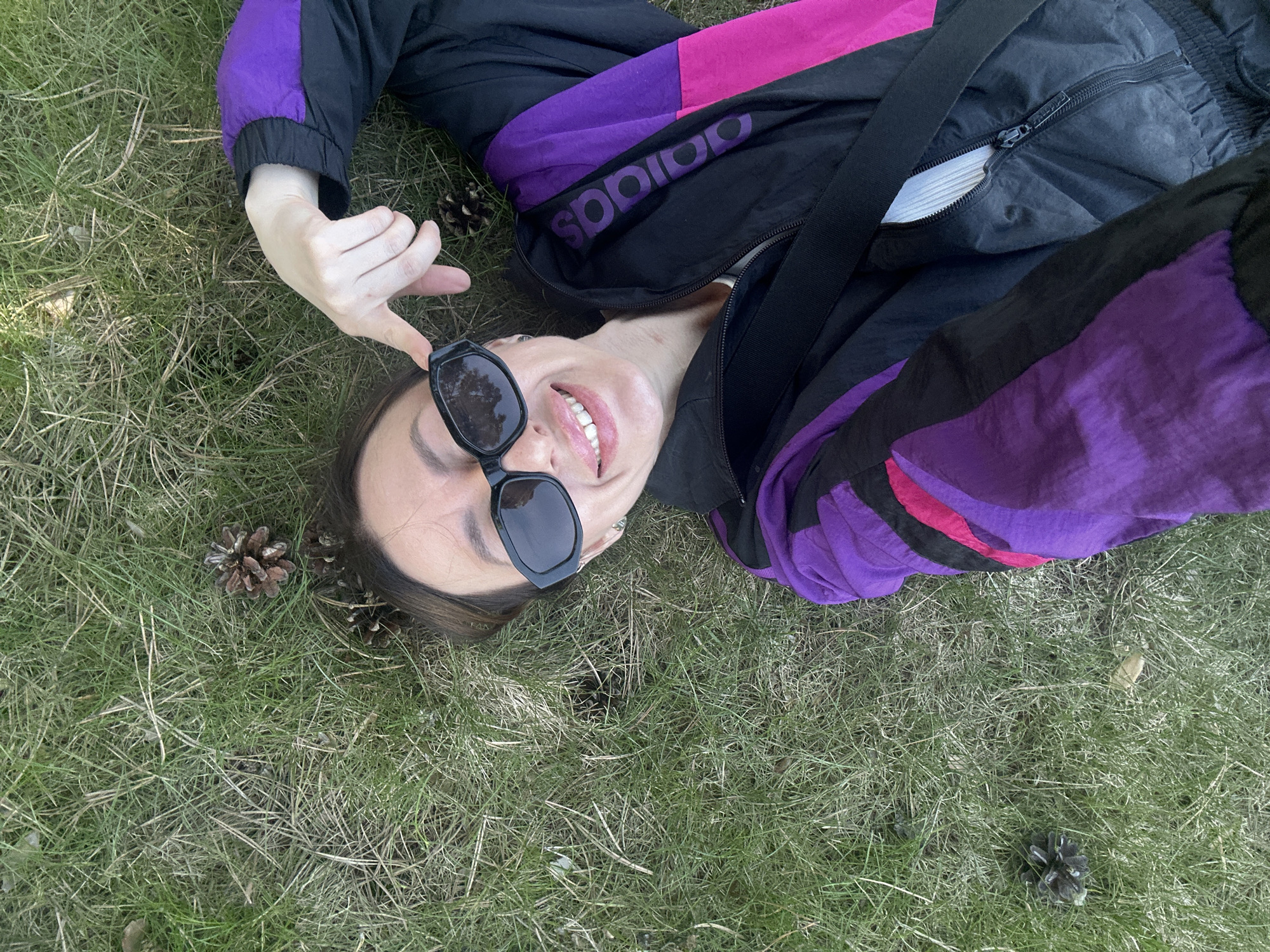 The image shows a person lying on a grass-covered surface with pine cones scattered around. The person is wearing sunglasses and a black Adidas tracksuit with purple and pink color blocking. They are smiling and pointing at their sunglasses with one hand. The location is unknown.