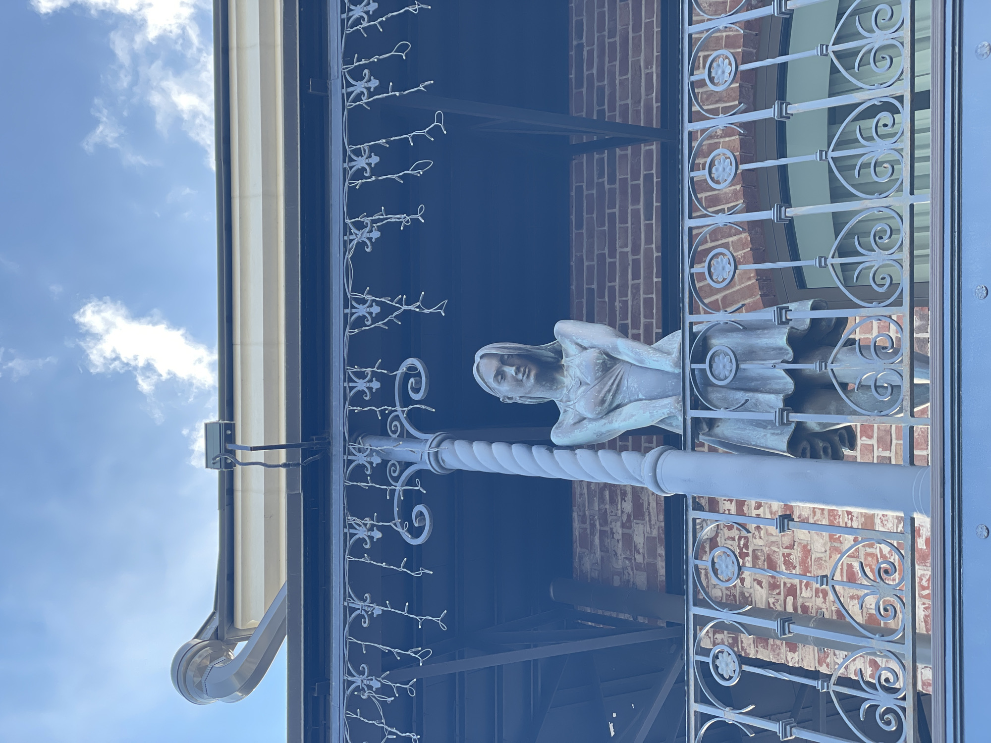 A low-angle shot depicts a pale, metal sculpture of a reclining woman with hands clasped near her chest. The sculpture is positioned on top of a support pole that appears to be part of an elaborate metal balcony railing featuring curly designs and flower-like motifs. Behind the railing, red brick walls can be seen. A dark, shaded area is visible under the balcony, with metal supports and beams. At the top left of the frame, a partially visible awning extends from the building. The sky, with scattered clouds, is visible in the upper left corner. The location is Kazan, Russia.