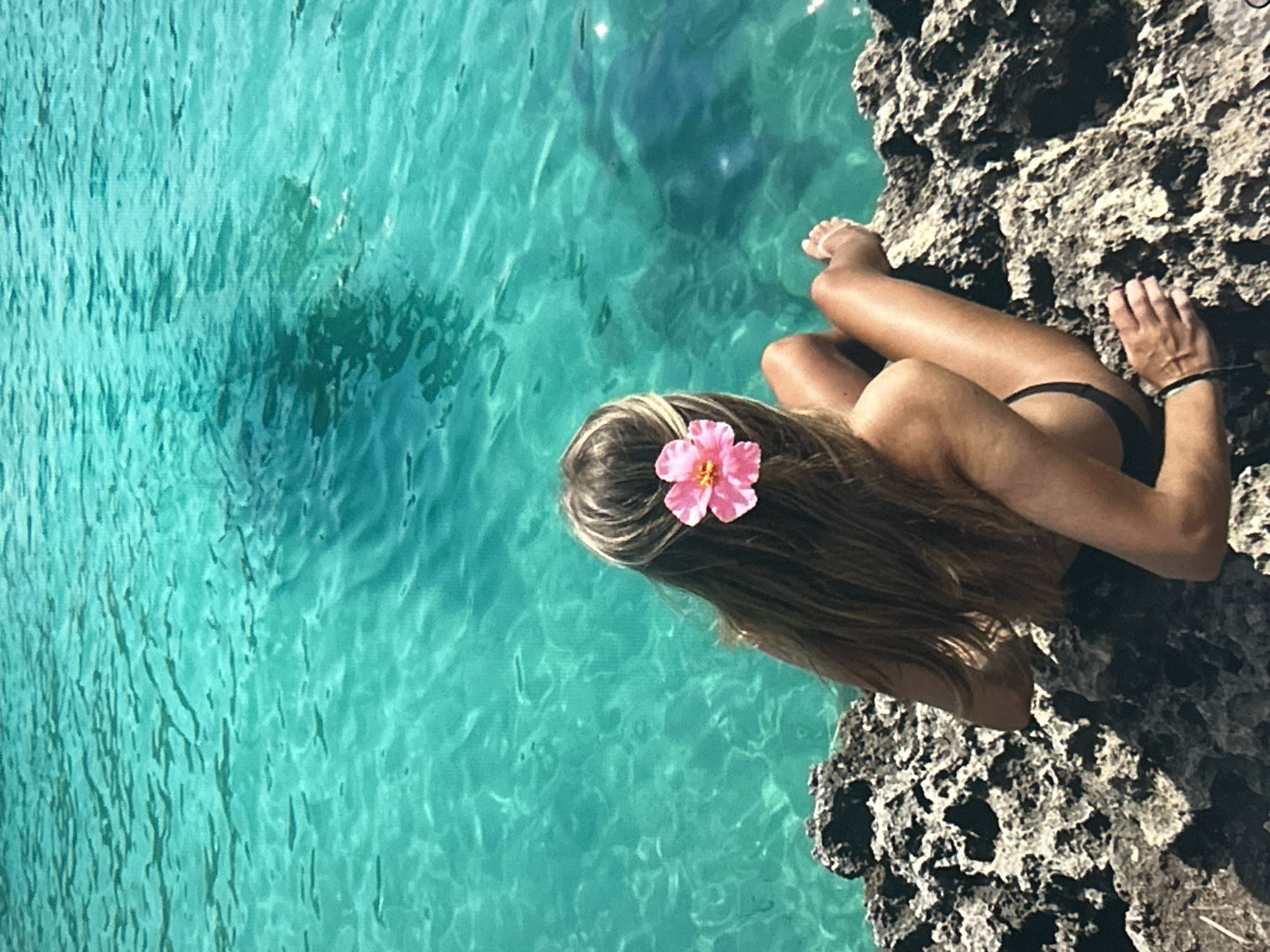 A high-angle, slightly rotated image captures a person reclining on a rocky outcrop beside clear, turquoise water. The person, viewed from behind, has long, light brown hair adorned with a single vibrant pink flower on the right side of their head. They are wearing dark swimwear, with tanned skin visible on their back, arms, and legs. Their right hand rests on the rocky surface, and their left arm is mostly out of view.
The water, which dominates the left and central parts of the frame, is strikingly clear, revealing the textured surface of the seabed or underwater formations below. Ripples and reflections are visible on the water's surface, indicating gentle movement. A large, dark, somewhat indistinct shape is visible submerged in the upper-center portion of the water, possibly a swimmer, diver, or a significant rock formation under the surface.
The rocky outcrop where the person is positioned is dark and craggy, providing a strong textural contrast to the smooth water. The scene suggests a sunny day in a tropical or pristine coastal location, emphasizing tranquility and natural beauty. There is no visible text in the image.