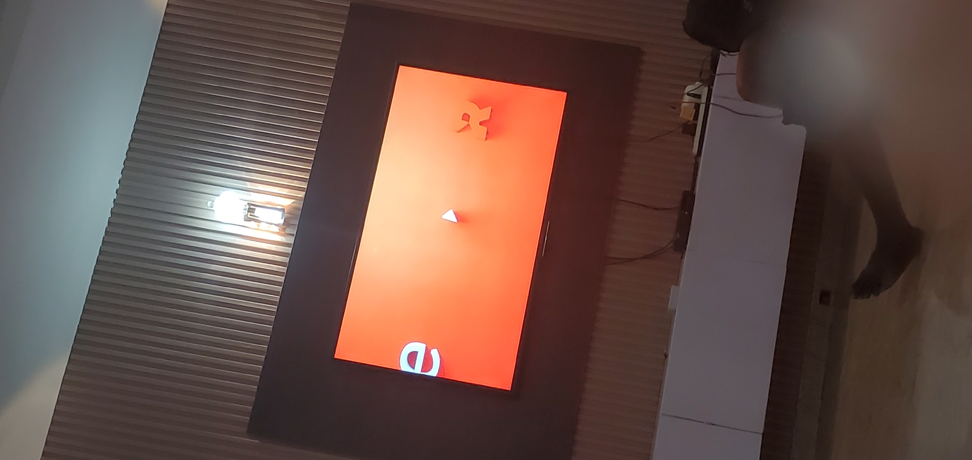 The image depicts an indoor scene, possibly a living room, with a large television screen displaying an orange background with two stylized characters: a white letter "e" on the left and an orange letter "R" on the right. A small white play button icon is centered on the screen. The television is mounted on a dark frame, which is set against a wall with vertical wooden paneling. A light fixture with a bright bulb is visible on the wall above and to the right of the television. Below the television, a white cabinet or entertainment center is present, with some electronic devices and cables on top of it. In the foreground, a person's leg and foot are visible on a wooden floor. The overall lighting suggests the room is dimly lit, with the television screen and the overhead light being the primary sources of illumination. The environment appears to be domestic.