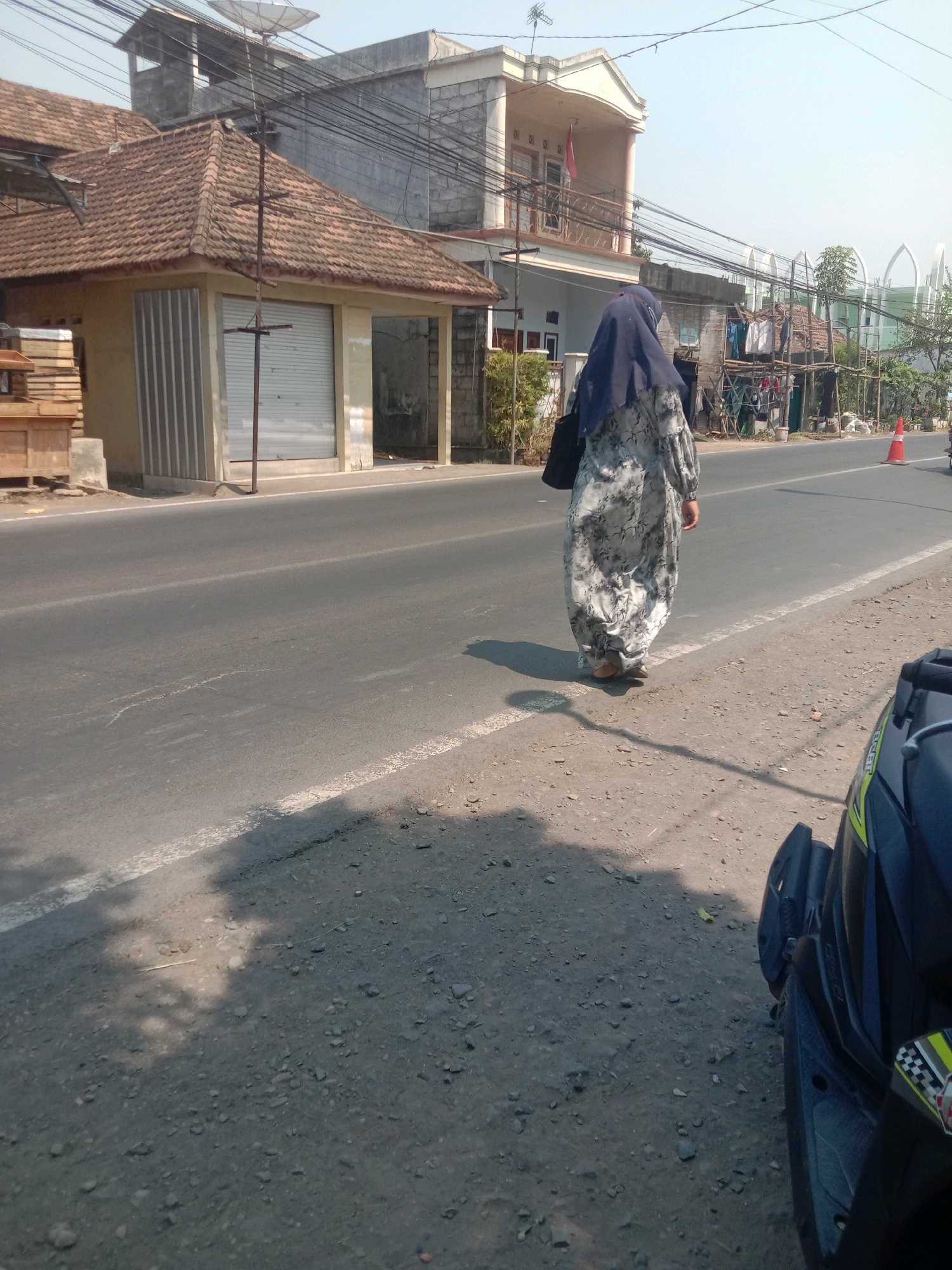 A woman wearing a dark blue hijab and a gray and white patterned dress walks away from the camera on the right side of a two-lane asphalt road. She carries a black bag over her left shoulder. On the left side of the road is a building with a tiled roof, a gray metal roll-up door, and a light yellow facade. Behind this building are more structures, including a two-story house with a balcony. Further down the road, on the right, there is a white and orange traffic cone and a structure with bamboo poles and some laundry hanging. The road surface is gray asphalt with a white line marking the edge. The ground on the side of the road is gravelly. The sky is clear and pale blue. A black motorcycle is partially visible in the lower right corner of the image. The scene appears to be in a residential or commercial area with buildings and a road.