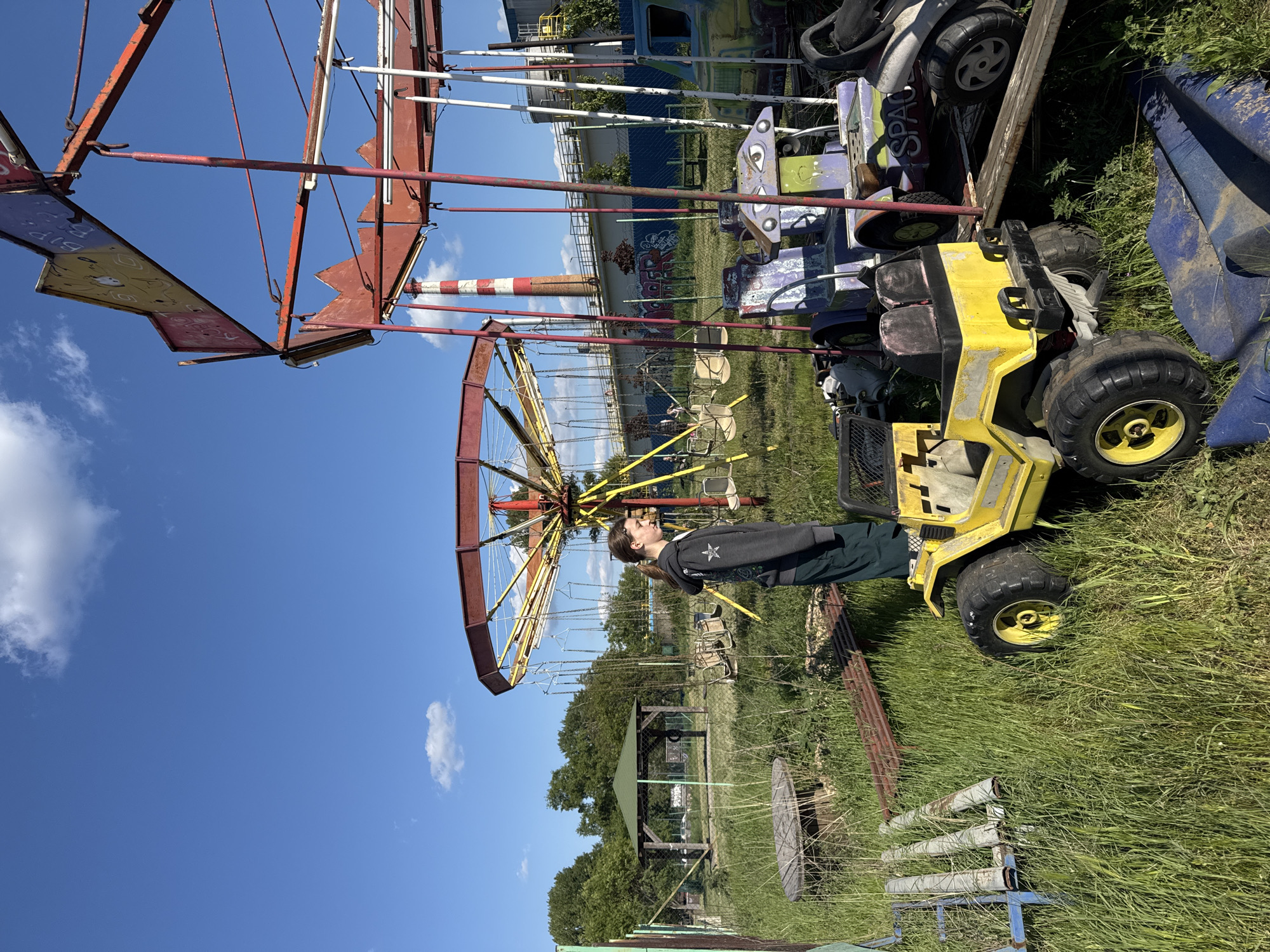 An outdoor scene captures a young person amidst a collection of abandoned amusement park rides and toy vehicles, set against a backdrop of overgrown grass, industrial structures, and a clear blue sky. The area appears neglected and disused.
A young person, likely a male teenager or young adult, with long brown hair, wearing a dark hoodie featuring a white star-like emblem and dark trousers, stands next to a yellow ride-on toy vehicle. The individual is looking towards the left, where a large, rusty fairground ride is prominently situated.
The fairground ride, possibly a swing carousel, is dilapidated, constructed from red and yellow metal framework with suspended chain seats. Near the person and to the right, several other faded and weathered toy vehicles are scattered, including the dominant yellow ATV-like ride and various smaller, colorful kiddie rides in purple, green, and blue hues. The partial word "SPACE" is visible on one of these toy vehicles, and "BIP" is partially visible on a panel of the larger fairground ride. The ground throughout the scene is covered in tall, green grass.
In the mid-distance, a wooden gazebo-like structure with a green roof is visible among trees. Further back, a tall, red and white striped chimney or stack stands out, along with other distant buildings and fences, hinting at an adjacent industrial or urban area.
The scene is brightly lit by daylight under a clear blue sky with a few scattered white clouds, indicating a sunny day. The overall impression is one of abandonment and decay within the Palilula Urban Municipality, Serbia, contrasting the vibrant natural light with the worn, disused equipment.